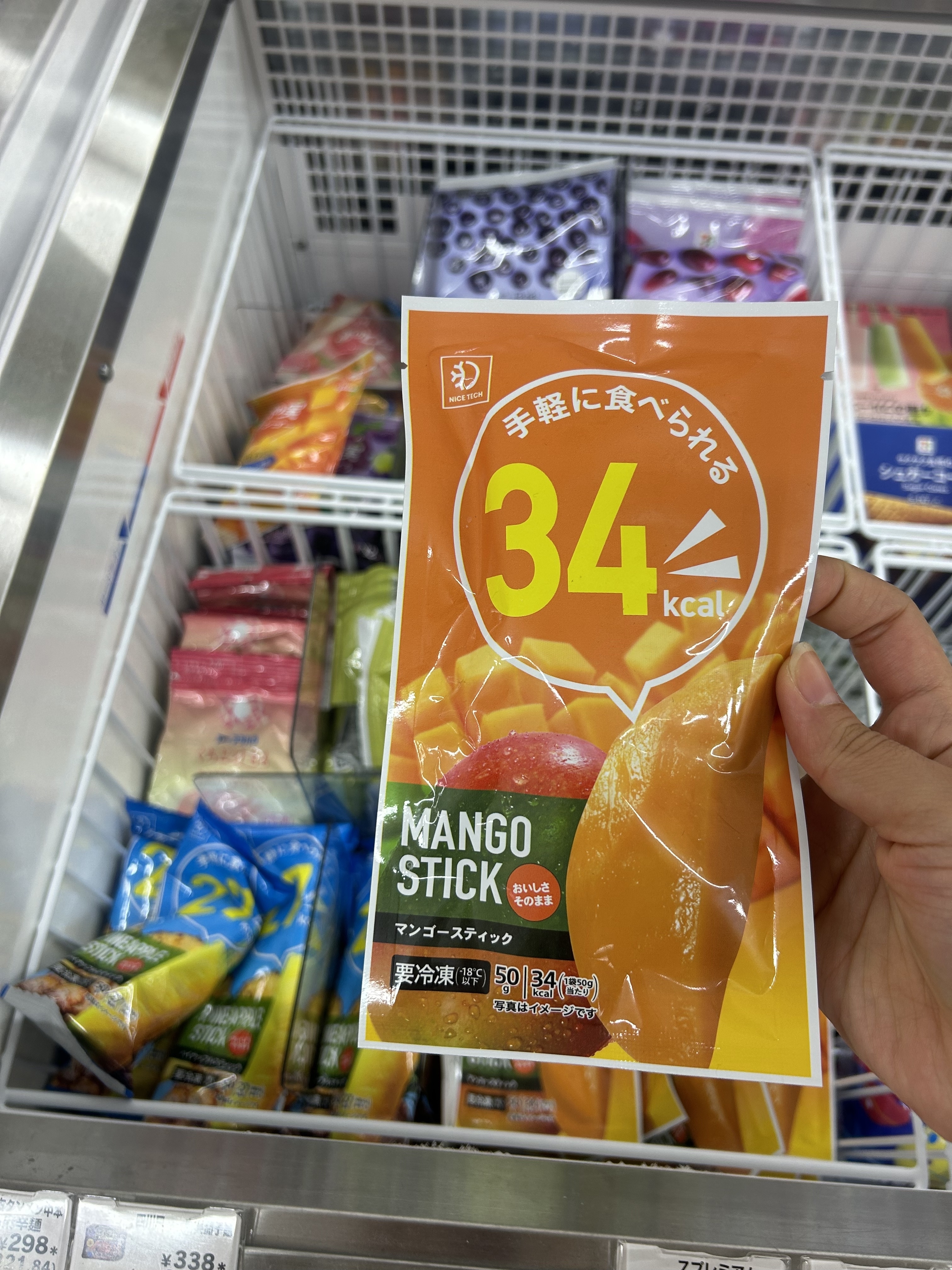A hand holds a package labeled "Mango Stick 34 kcal" in front of a freezer section filled with similar packaging and various frozen fruit snacks