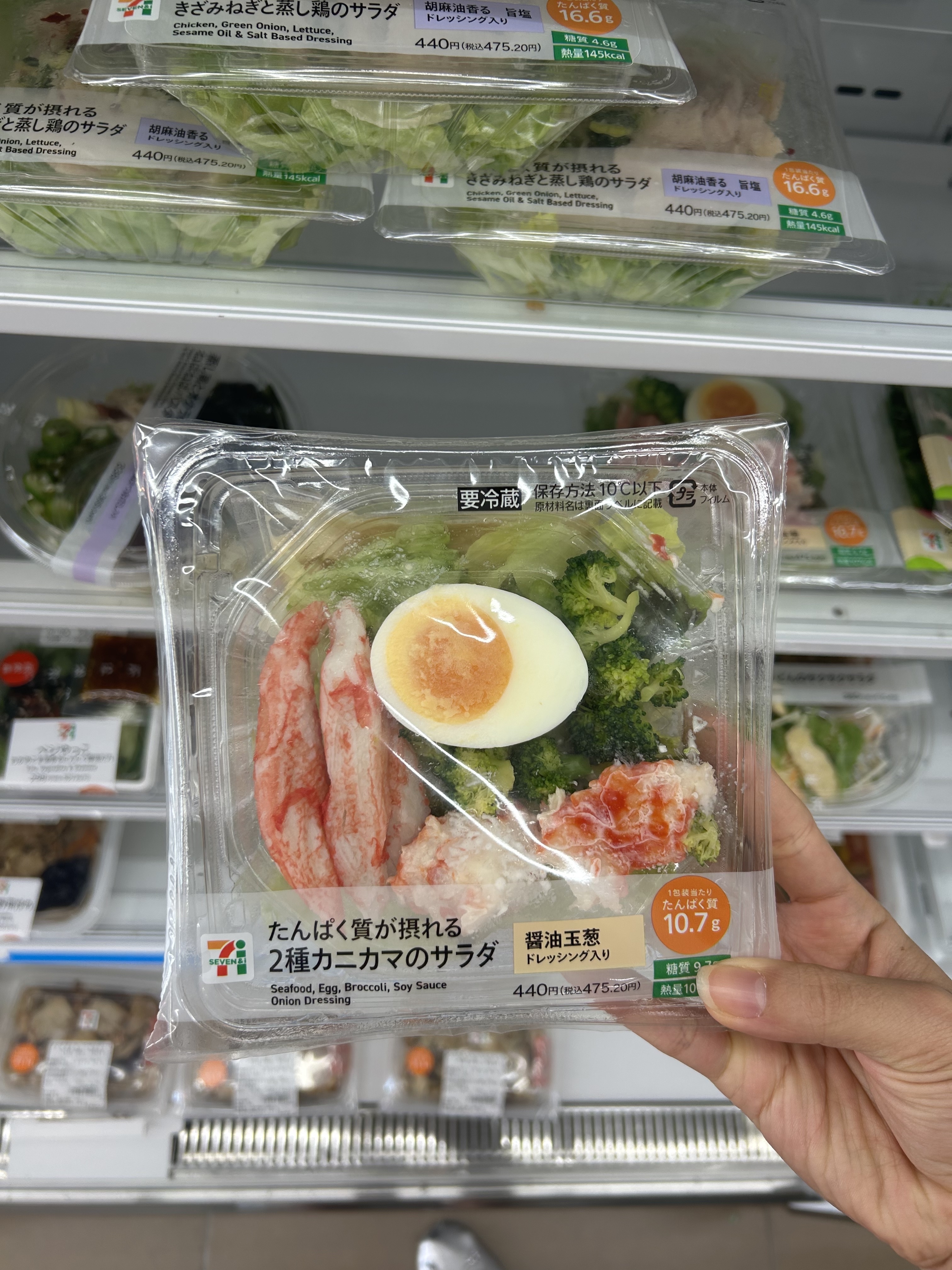 Hand holding a packet of 7-Eleven salad featuring imitation crab, greens, and a boiled egg. Shelves stocked with other salads are in the background