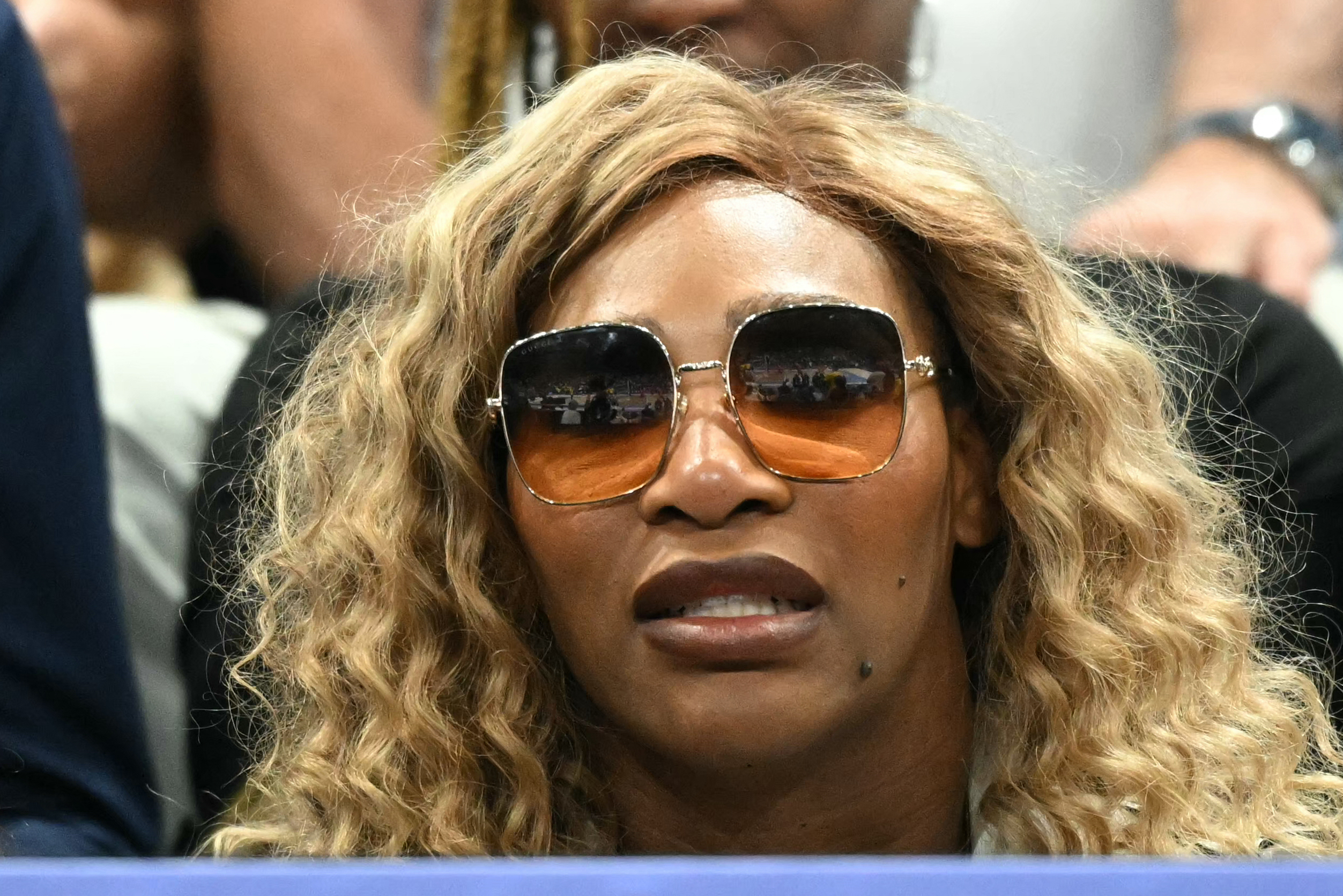 Serena Williams with wavy hair and sunglasses, watches from the audience