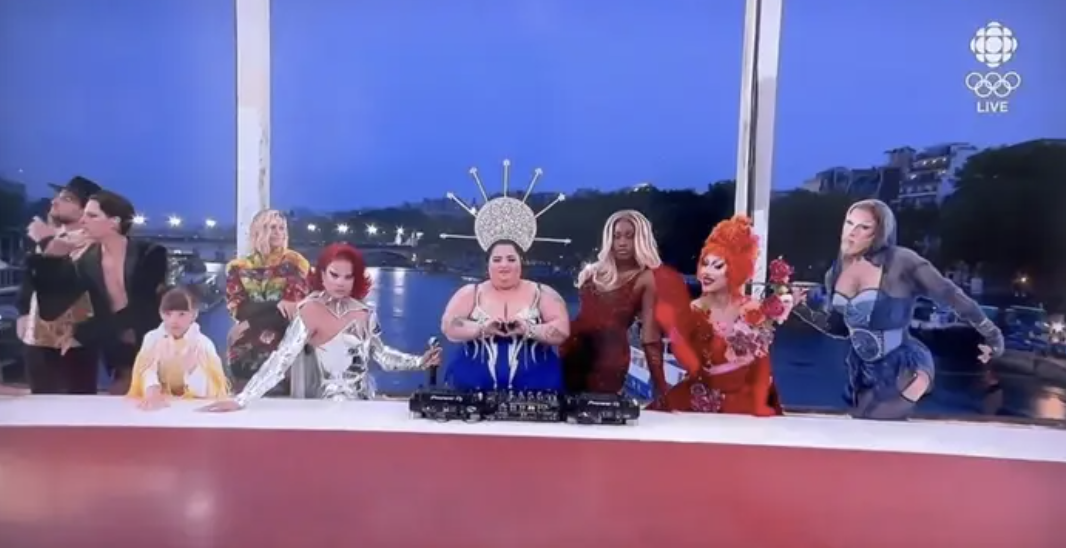 Lip-syncing performers on a live CBC broadcast featuring drag queen characters in vibrant costumes and headpieces, including DJ with spiked headpiece in center