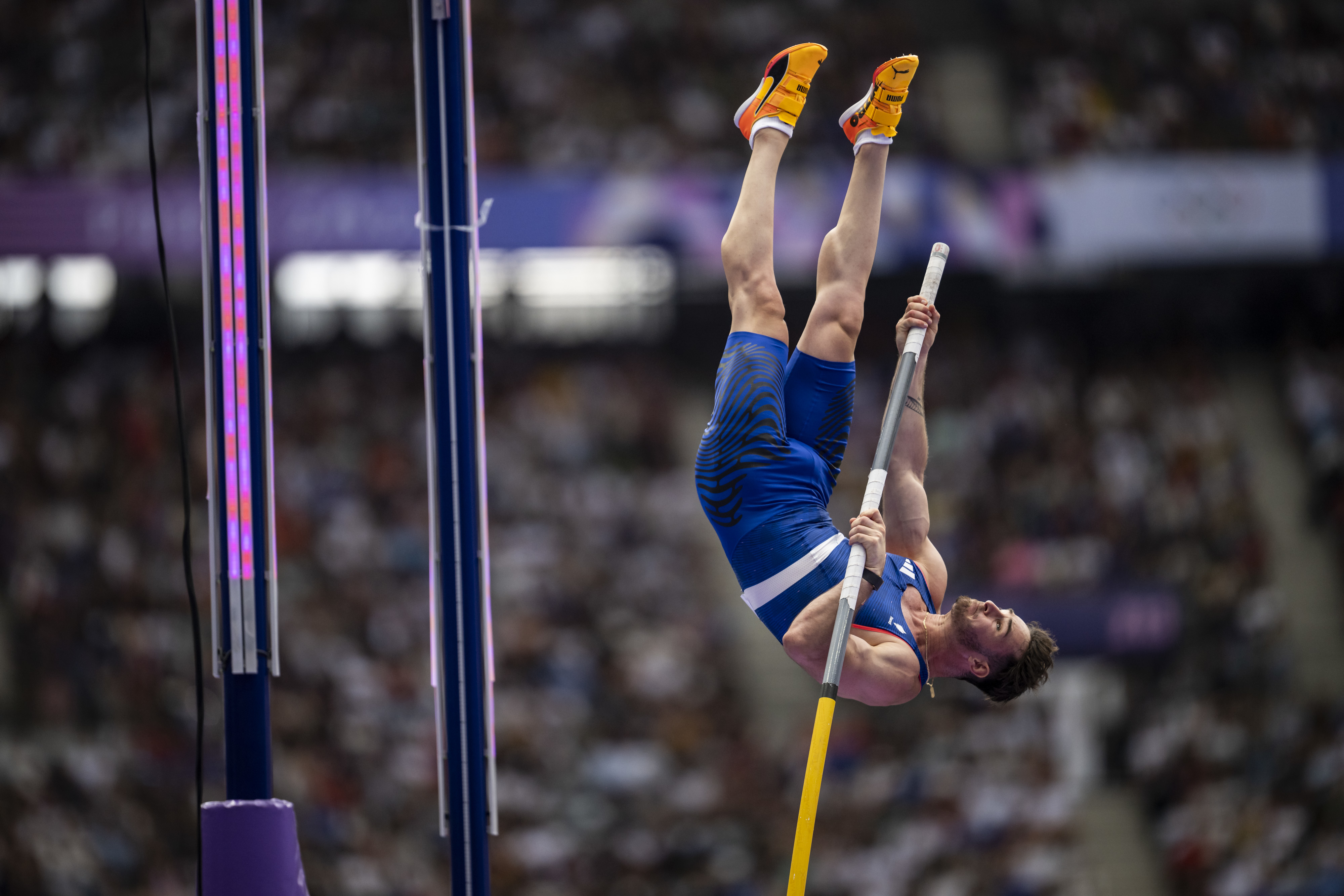 A pole vaulter wearing a blue athletic outfit and orange shoes is captured mid-air during a vault in a stadium filled with spectators