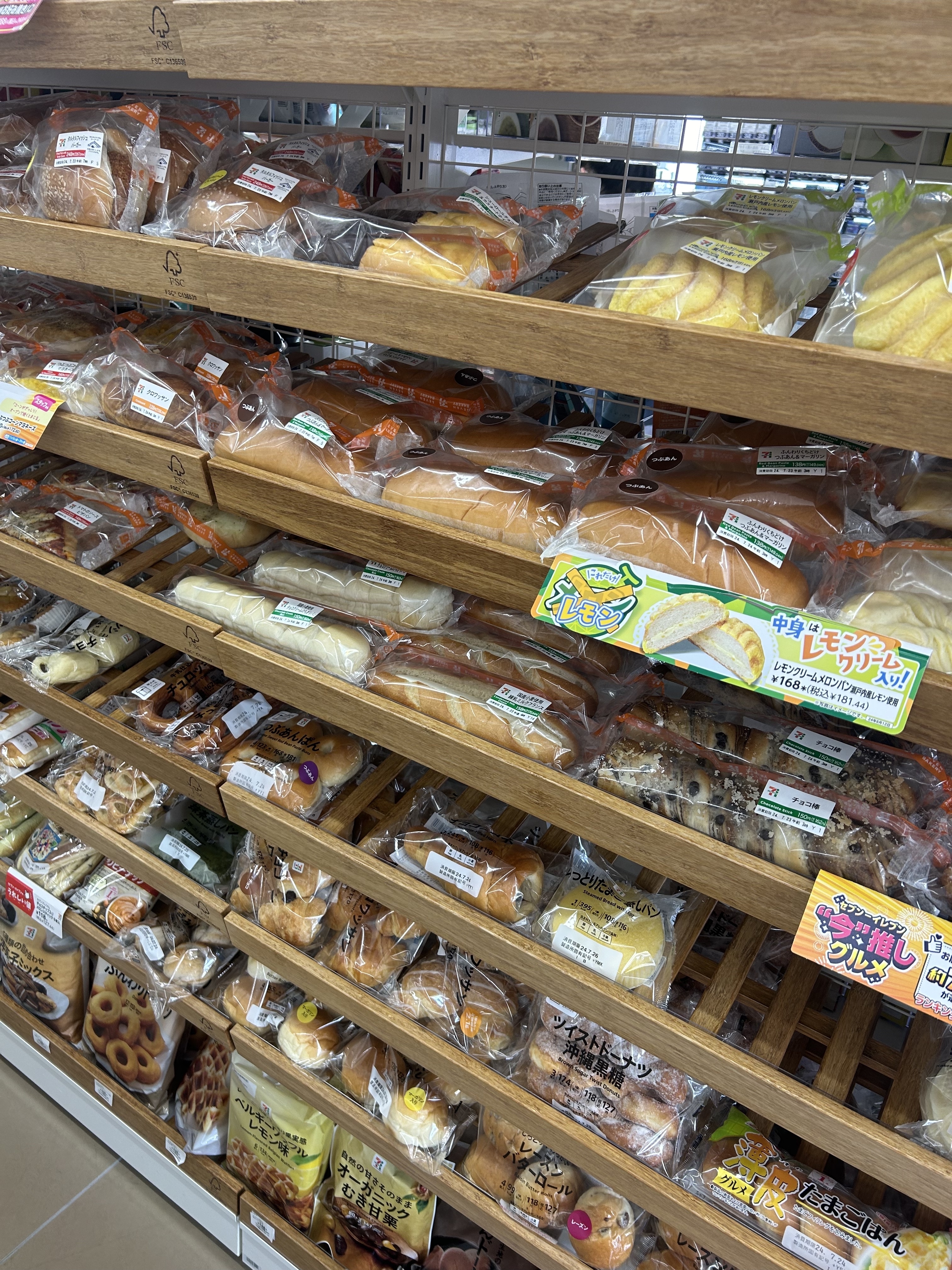 A bakery display with various packaged breads and pastries on wooden shelves, featuring items like sliced bread, rolls, muffins, and other baked goods