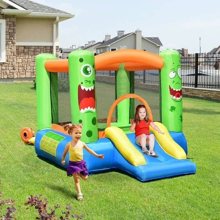 Two joyful children, one sliding and one running, play on a monster-themed inflatable bounce house in a backyard