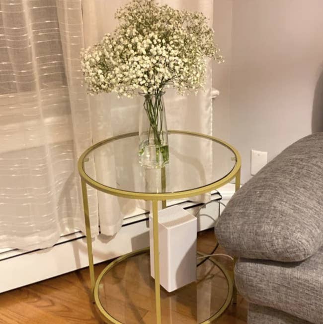 Glass side table with a small vase of baby's breath flowers, placed next to a grey sofa and sheer curtains