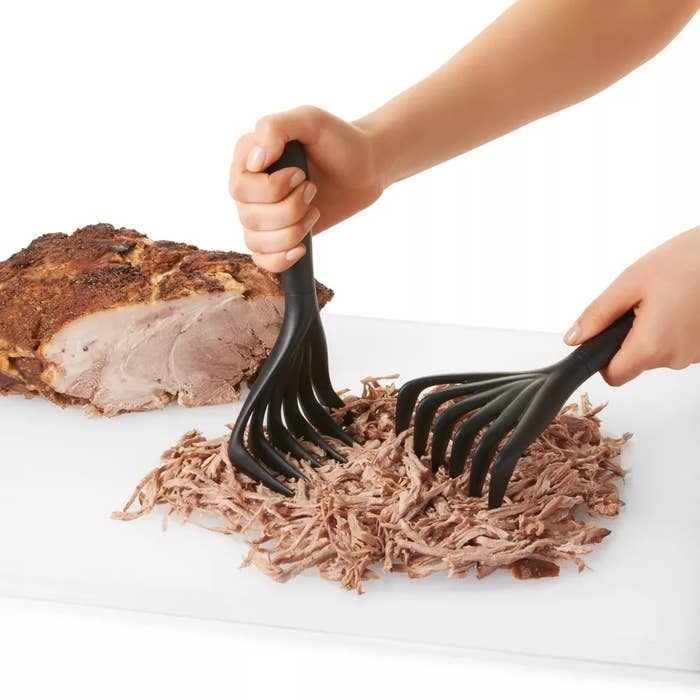 Hands using black shredding claws to shred cooked meat on a white cutting board, with a piece of unshredded meat nearby