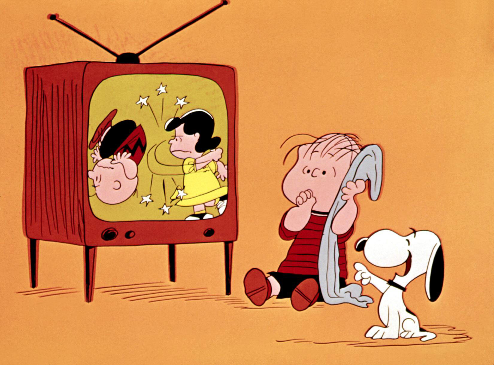 Charlie Brown gets kicked by Lucy on TV, while Linus is holding his blanket and Snoopy watch