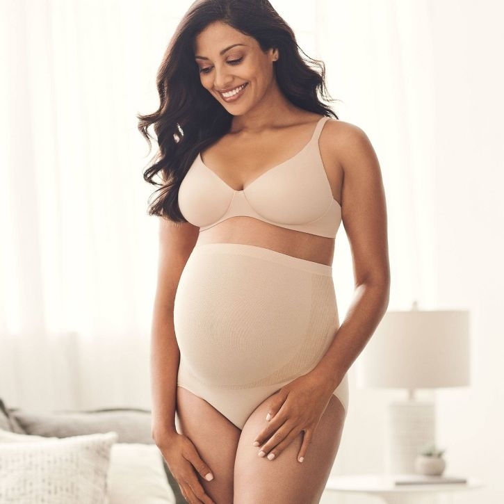 Pregnant woman poses in maternity shapewear set, smiling and cradling her belly
