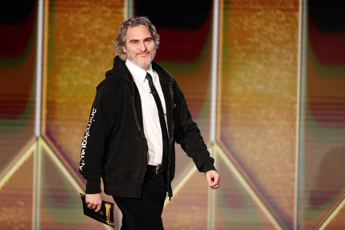 Joaquin Phoenix on stage wearing a shirt, tie, and hoodie, holding an award envelope and smiling