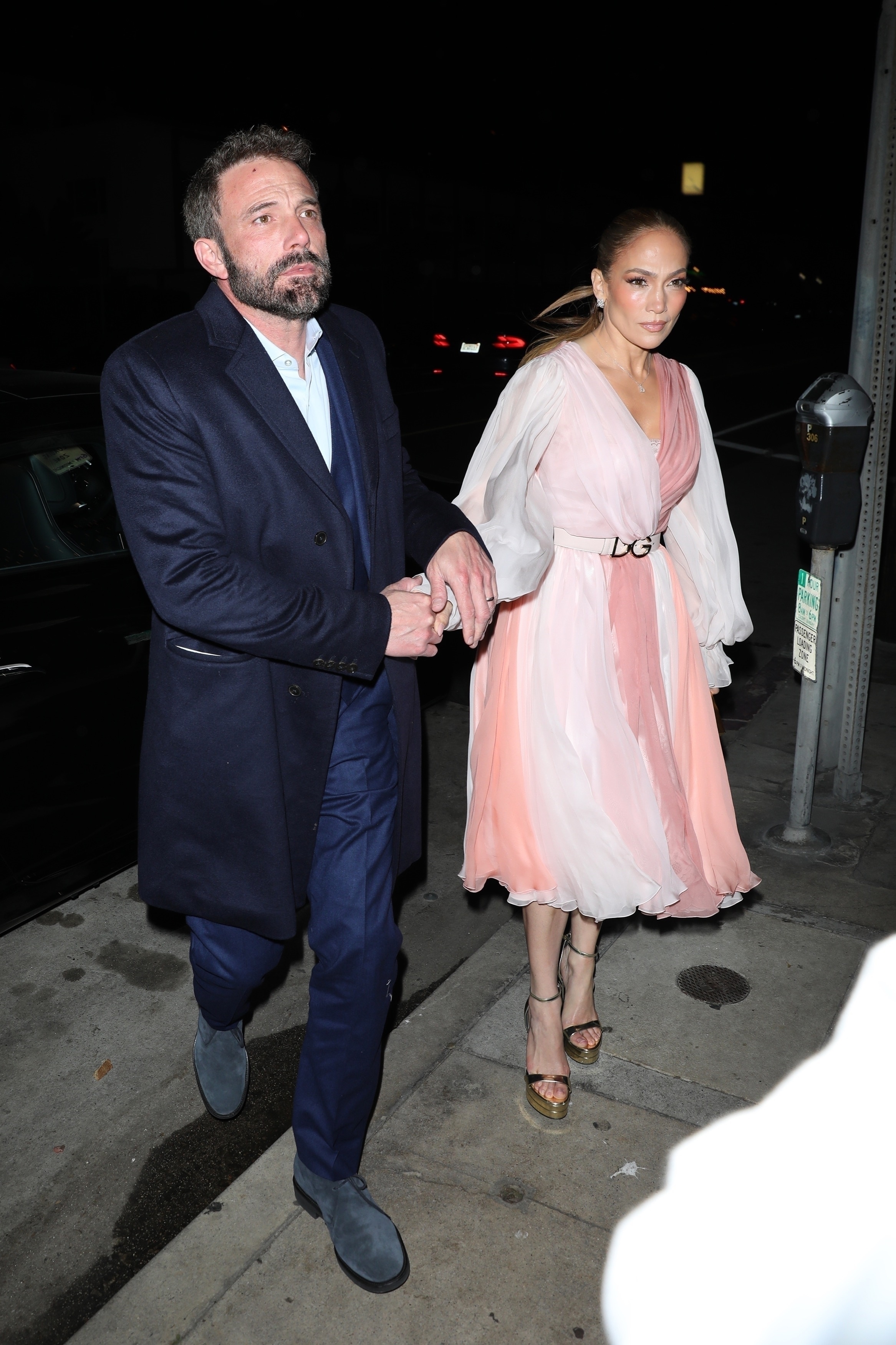 Ben Affleck and Jennifer Lopez holding hands, walking on a street at night. Ben is in a formal suit; Jennifer is in a stylish flowing dress and heels