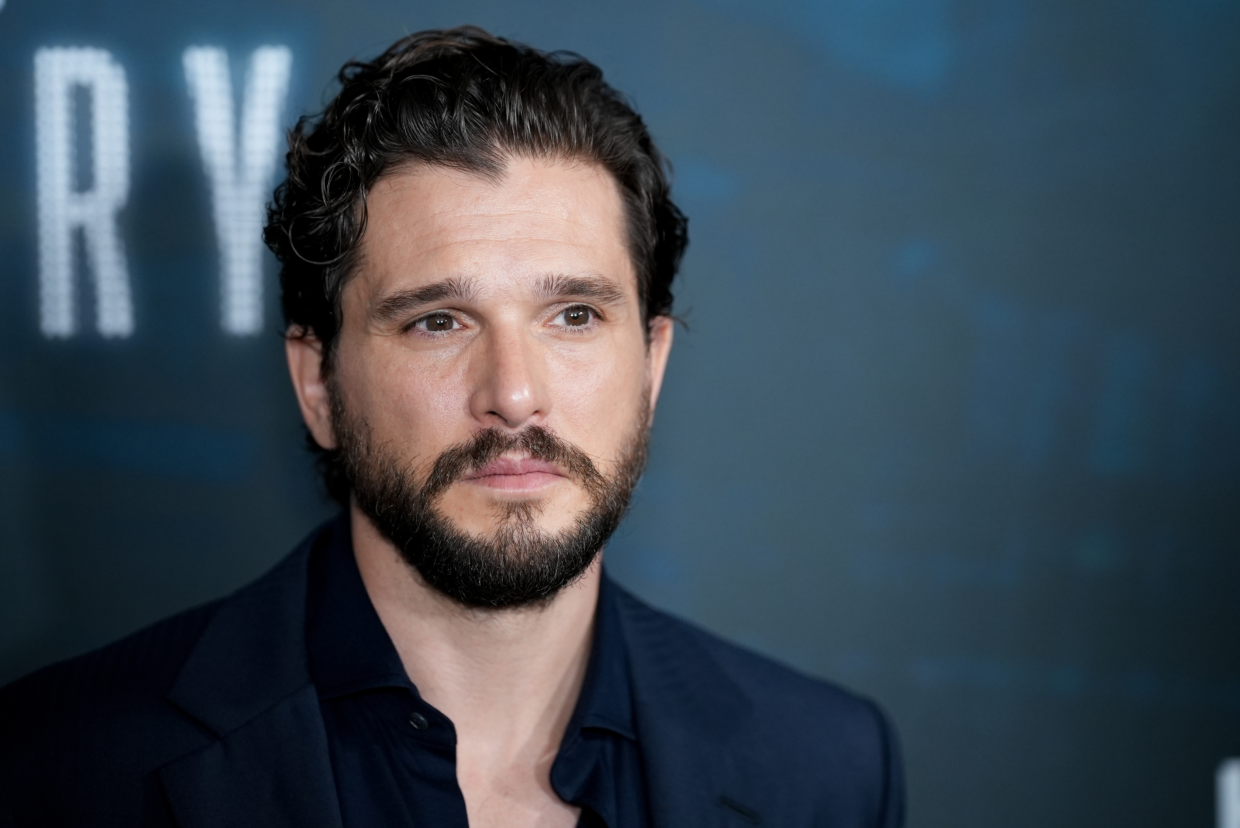 Kit Harington at an event, wearing a dark suit and looking into the distance with a serious expression