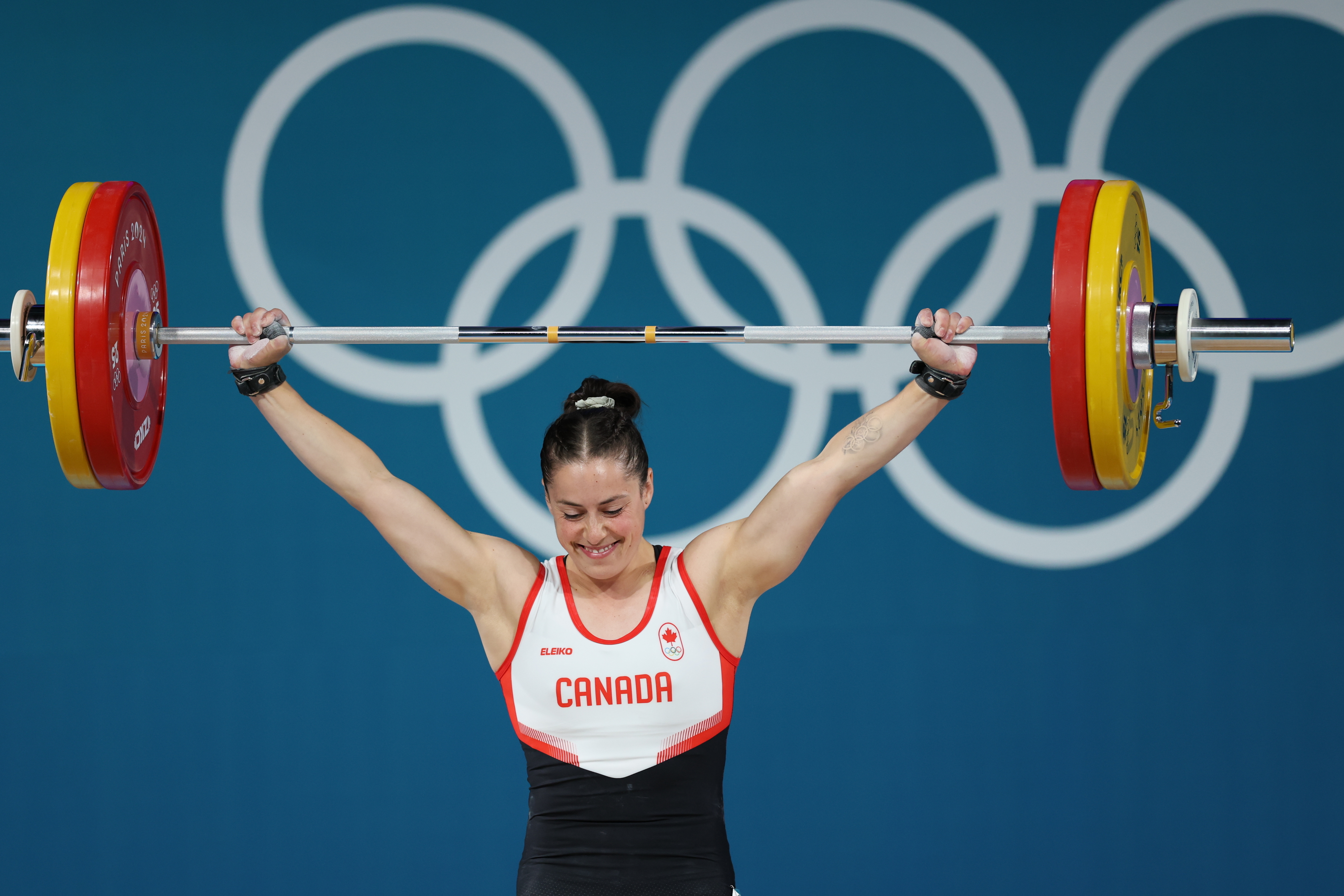 Weightlifter wearing a "Canada" uniform lifts a barbell with Olympic rings in the background