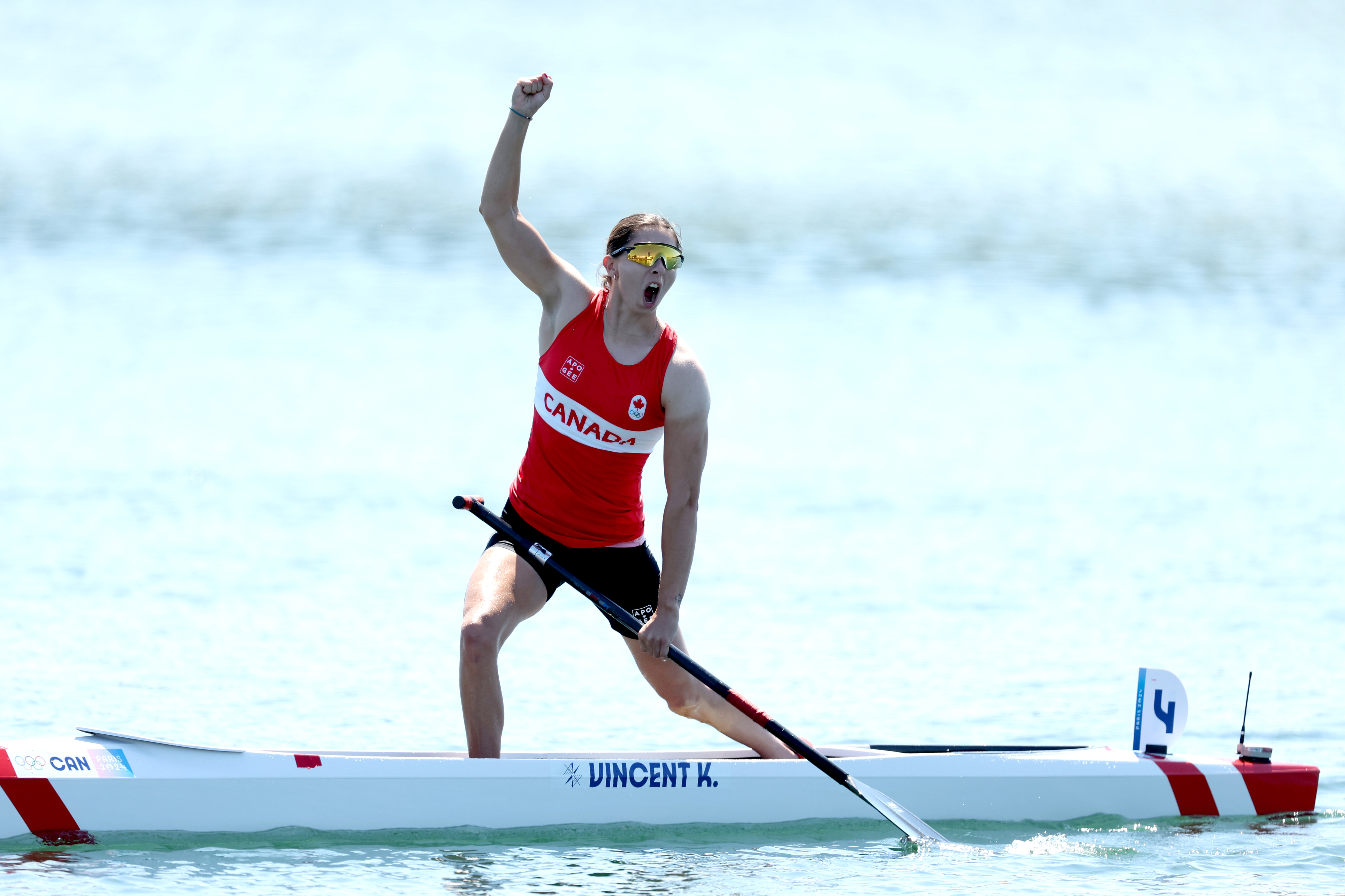 Vincent-Lapointe celebrates after a canoe race, wearing a 'Canada' sports shirt and holding a paddle while standing in her canoe on the water