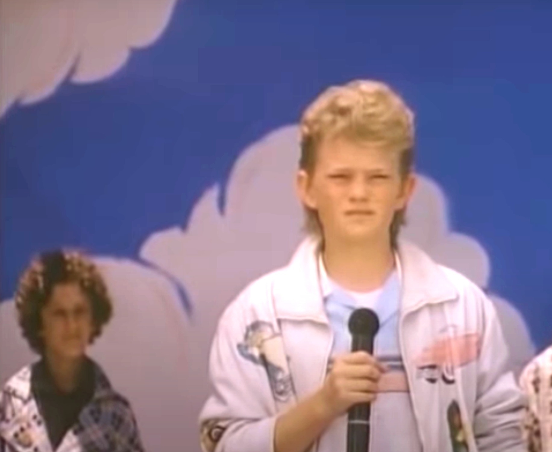 A young Neil grips a microphone, standing in front of a painted backdrop with clouds. young Dustin with curly hair stands in the background