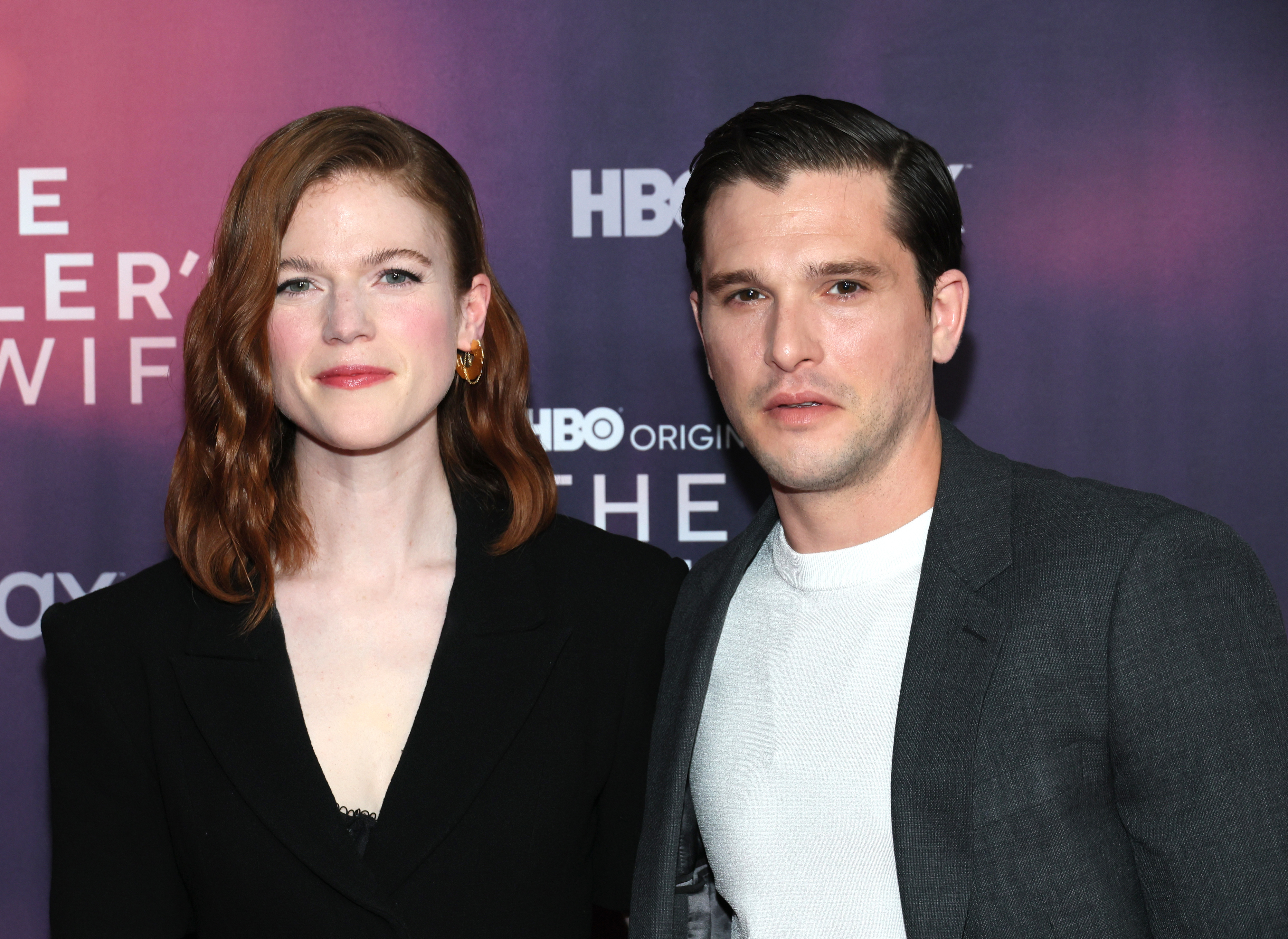 Rose Leslie and Kit Harington pose against a backdrop with HBO max logos at a public event. Rose wears a black suit, and Kit dons a grey blazer over a white shirt