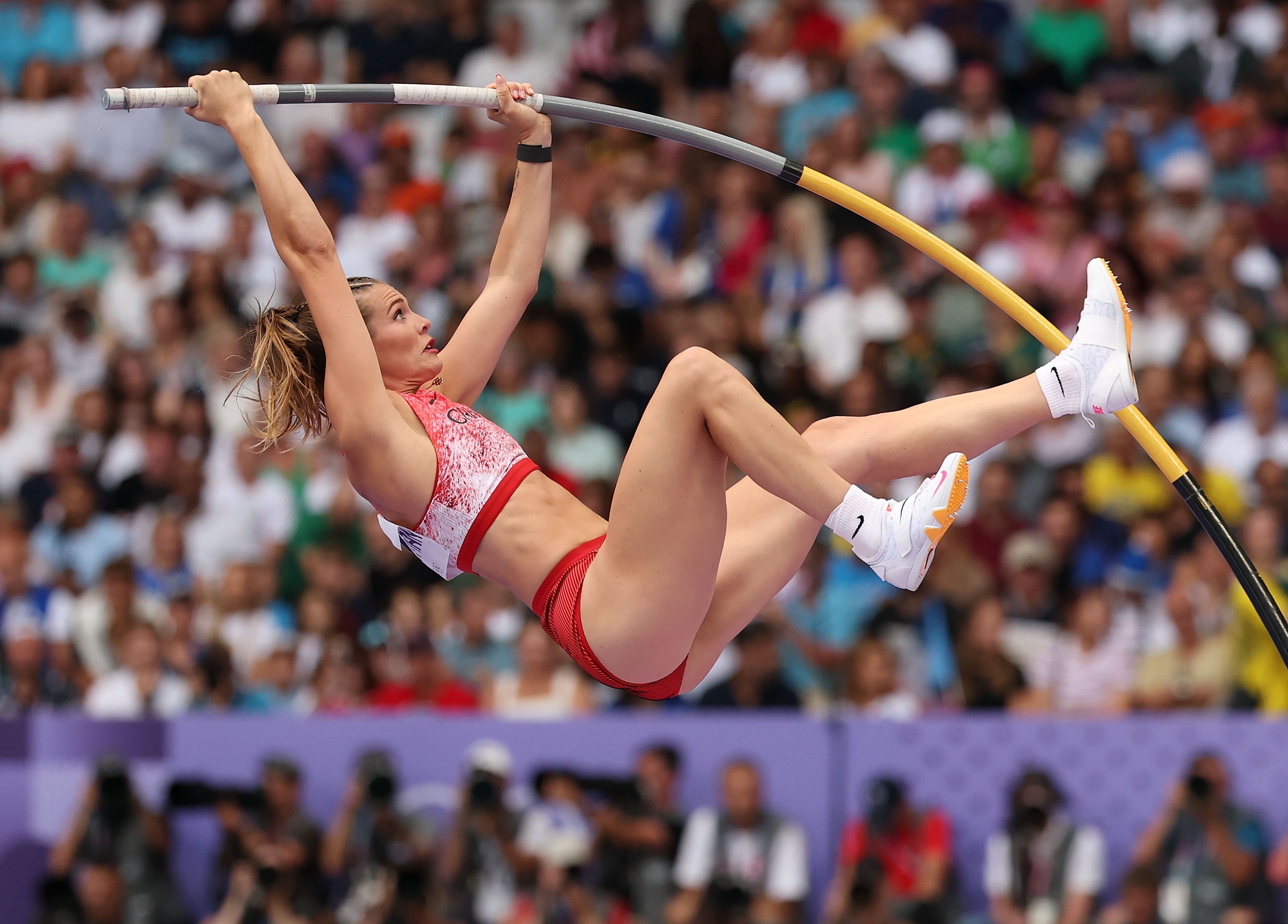 Pole vaulter jumping during a sports event, wearing a crop top and shorts, and holding the pole with both hands. Crowd watches from the background