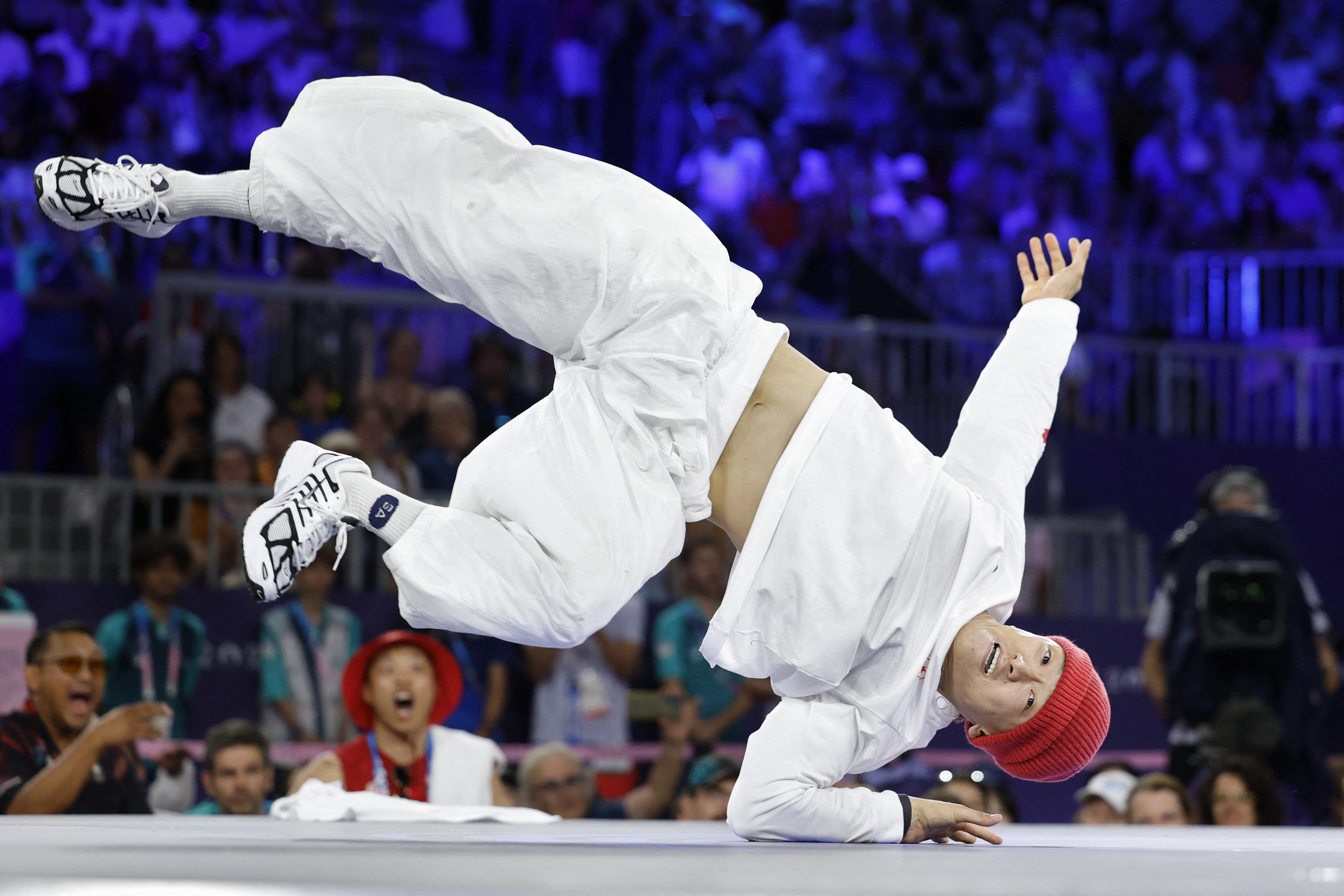 An athlete in loose white clothing and a red beanie performs a dynamic breakdancing move on the floor at a sporting event