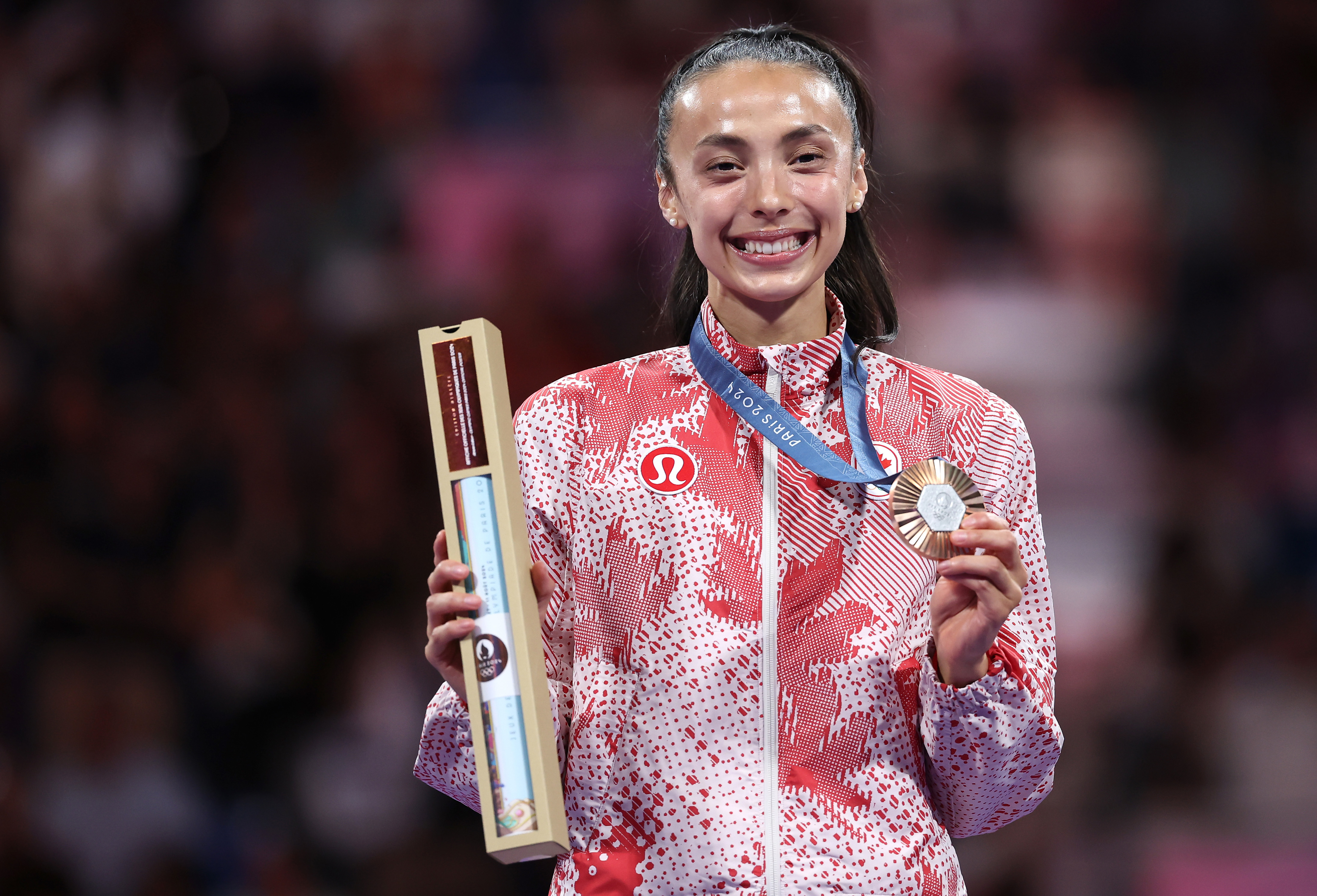 Athlete Rebeca Andrade smiles while holding a medal and a certificate during a sports event. She is dressed in sporty attire