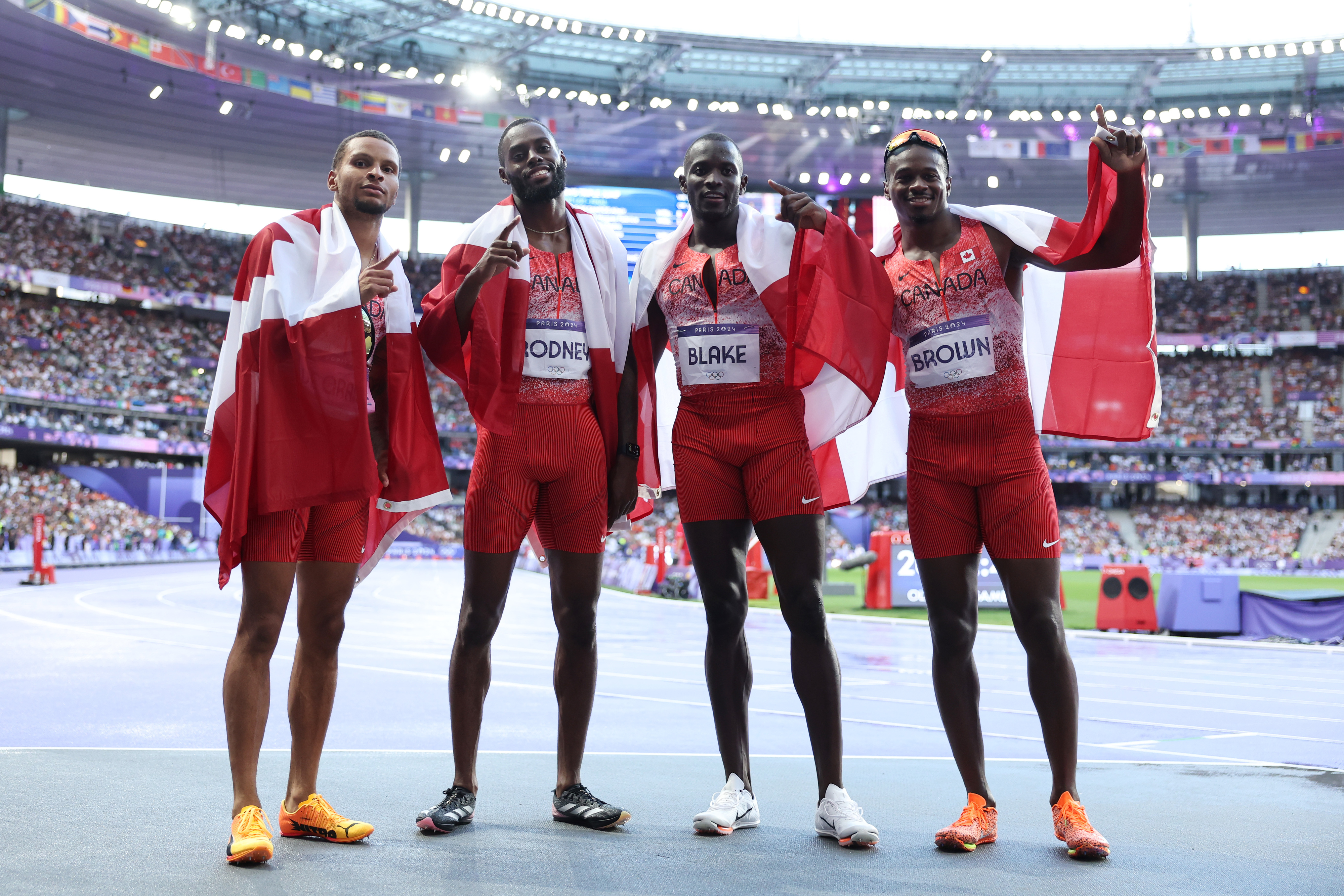 Athletes Brendon Rodney, Yohan Blake, Jerome Blake, and Aaron Brown pose on the track wrapped in flags, celebrating at a stadium
