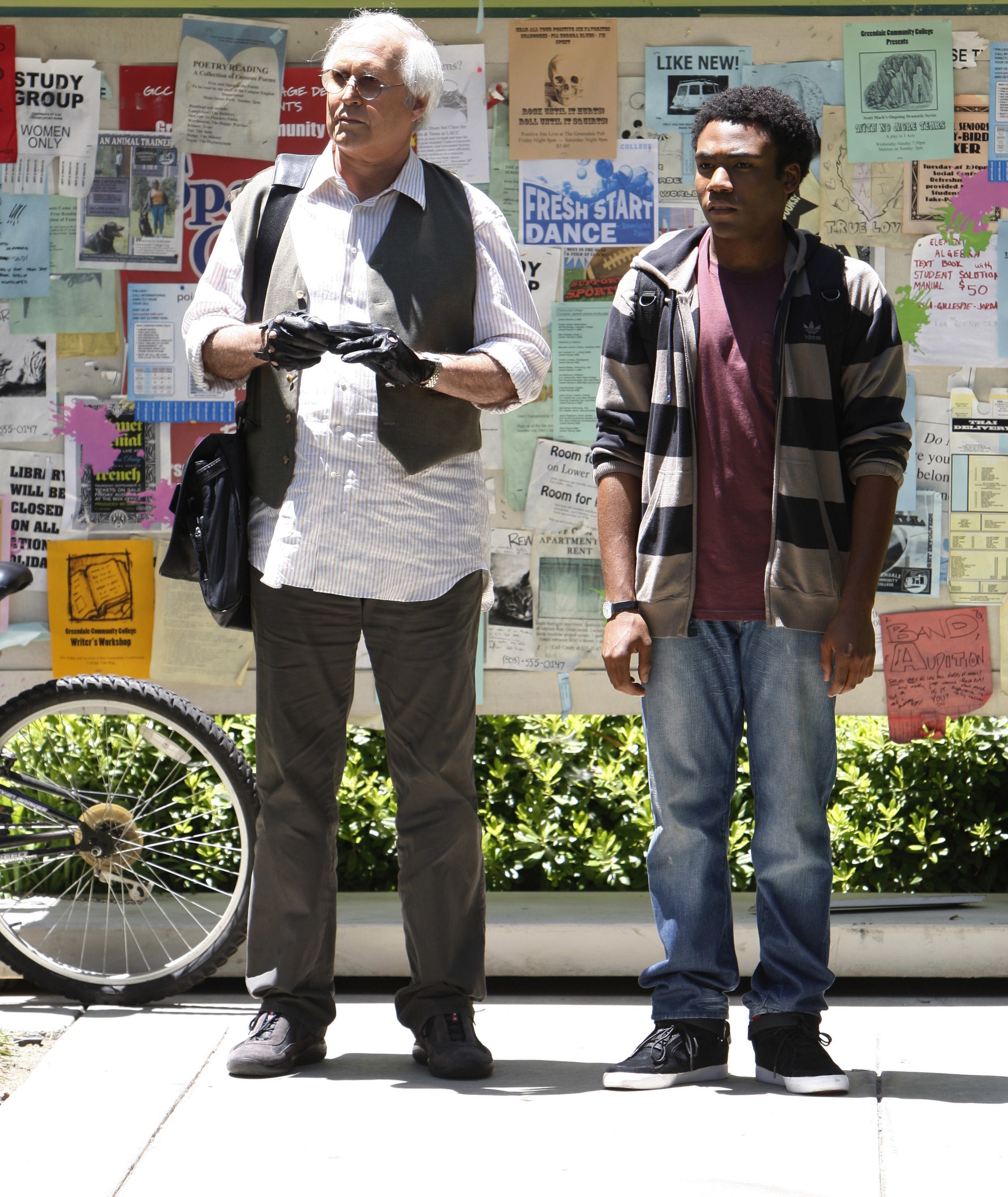 Chevy Chase and Donald Glover stand in front of a wall covered with flyers. Chevy wears a vest over a shirt, Donald in a striped hoodie and jeans