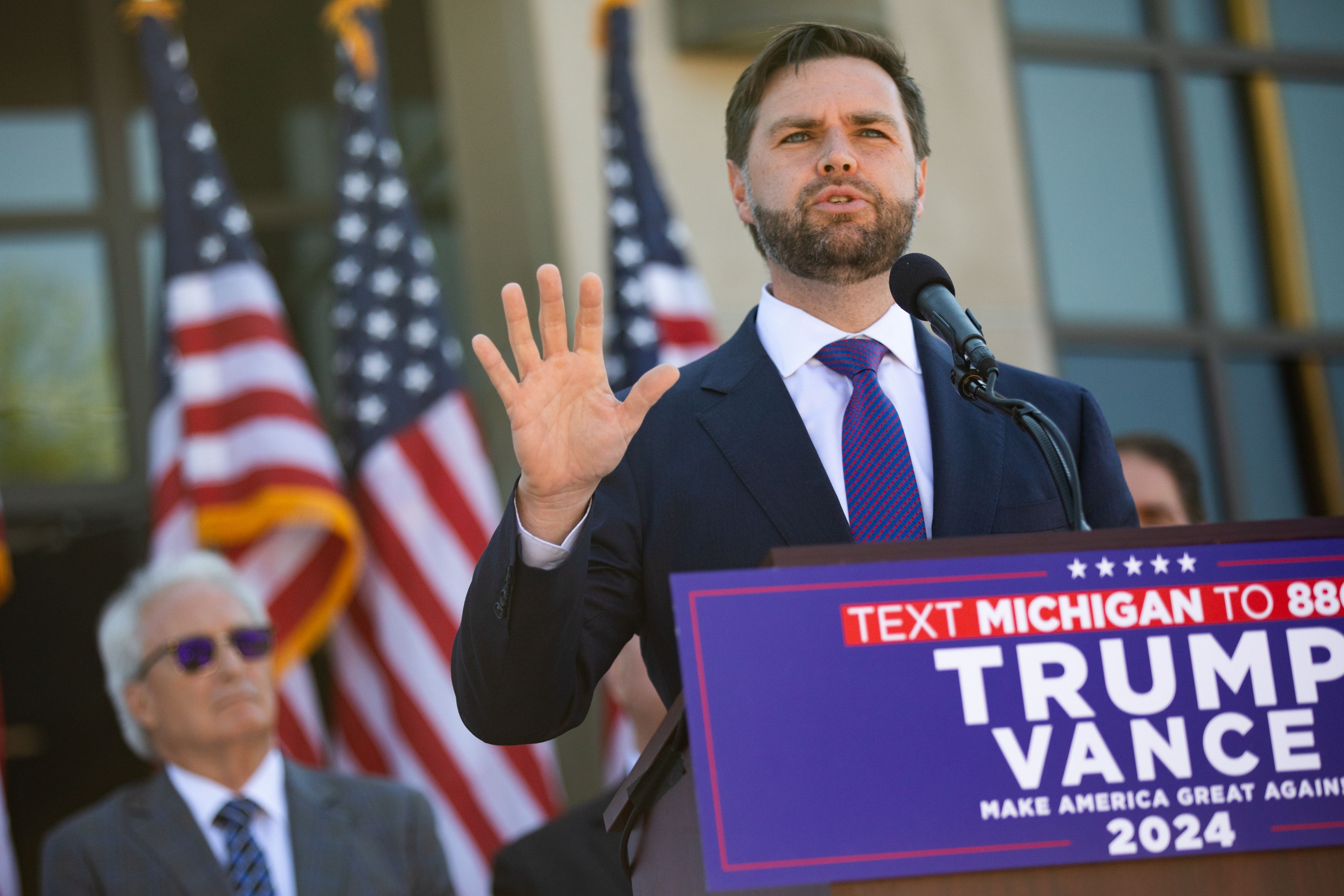 J.D. Vance speaking at a podium with a &quot;Text Michigan to 88022 Trump Vance Make America Great Again 2024&quot; sign, with American flags in the background