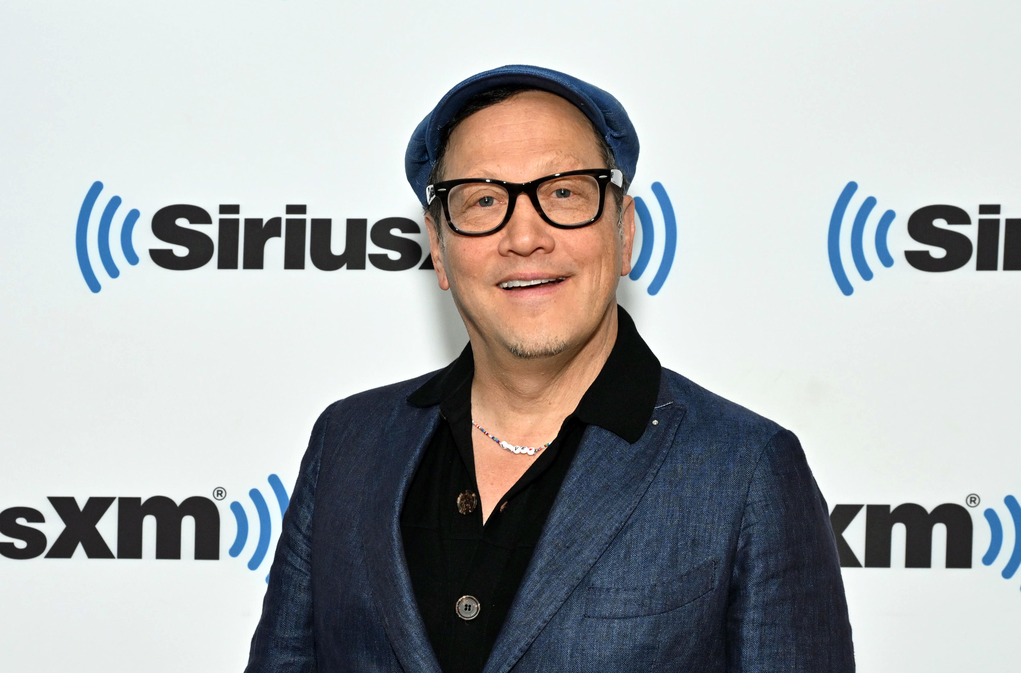 Rob Schneider in a casual blue blazer, black shirt, glasses, and a matching cap at a SiriusXM event backdrop
