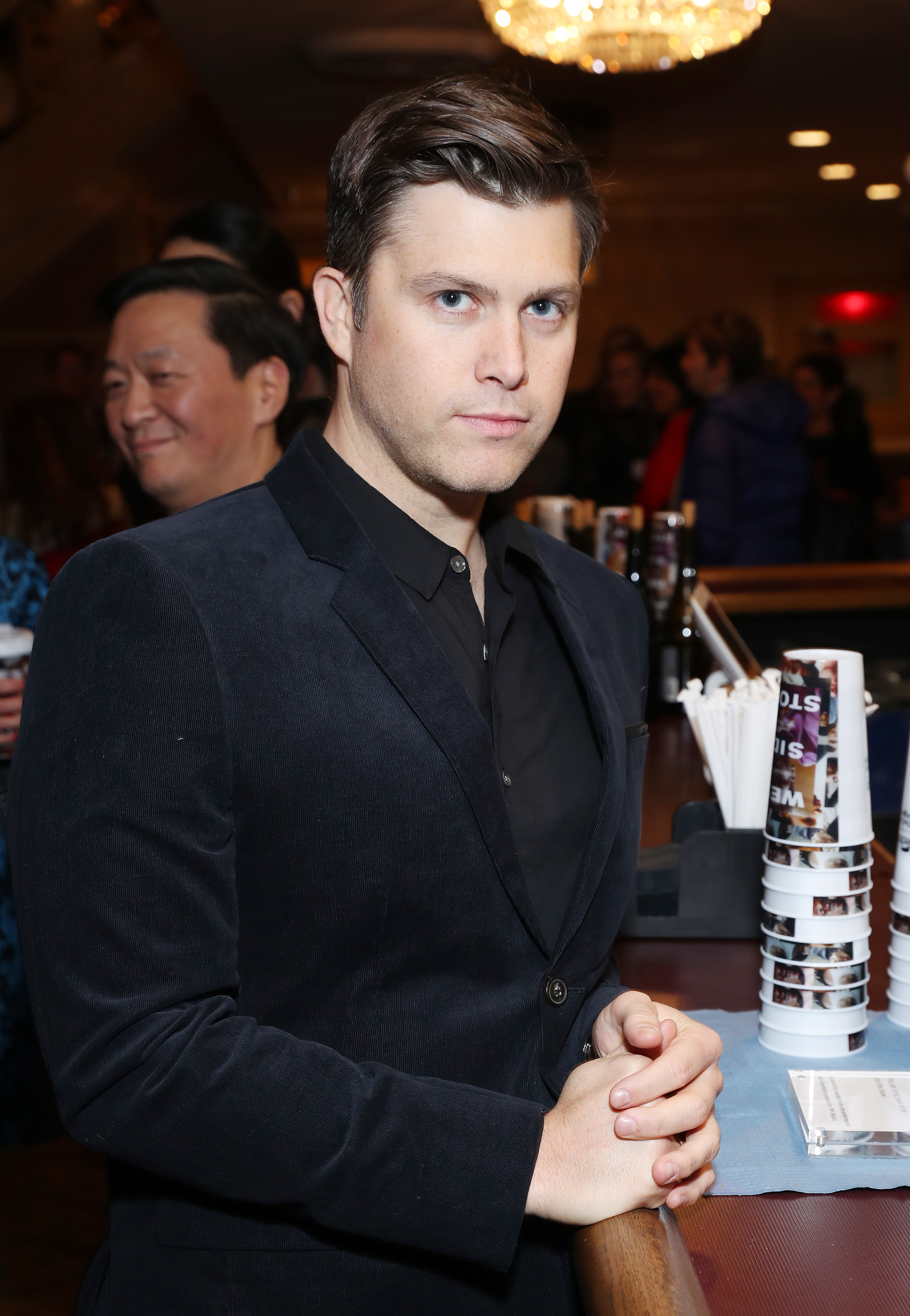 Colin Jost stands at a bar, wearing a dark suit and shirt, with hands clasped. In the background, others are socializing
