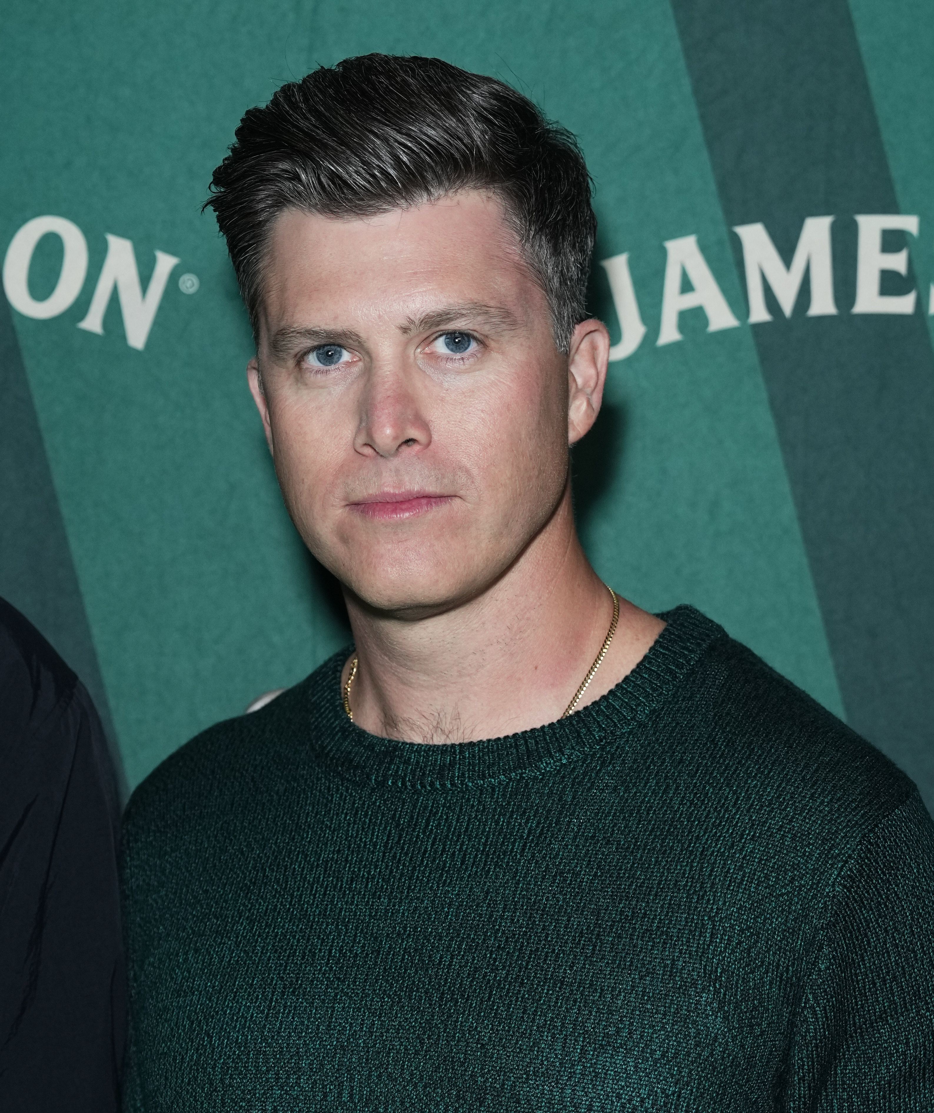 Colin Jost wearing a knit sweater stands in front of text that reads "Jameson."