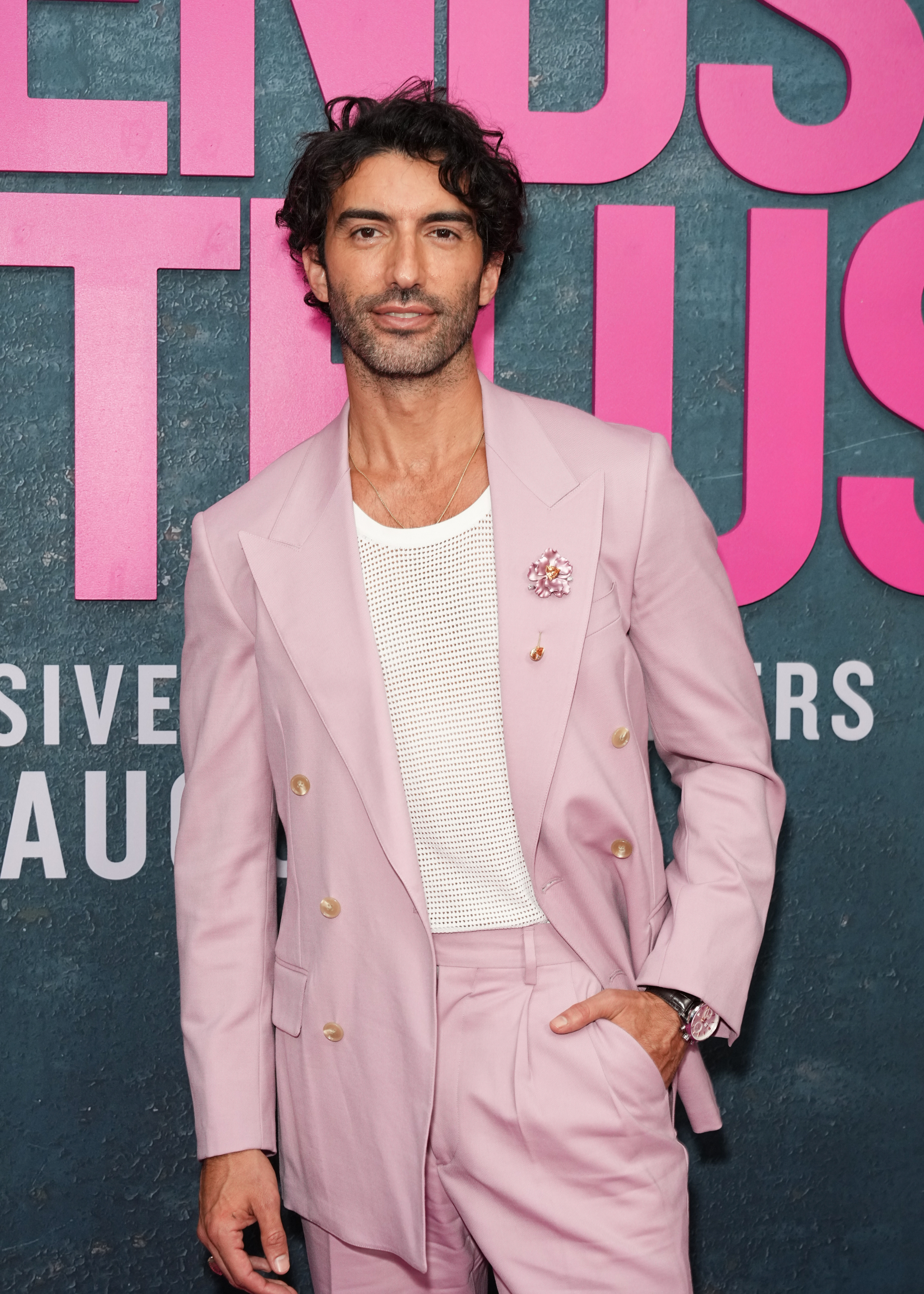 Justin Baldoni stands in front of a backdrop wearing a stylish pink suit and white mesh shirt at the &quot;Friends &amp;amp; Truth&quot; event