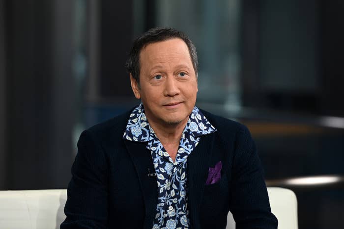 Rob Schneider is seated indoors, wearing a patterned shirt and a dark blazer with a pocket square