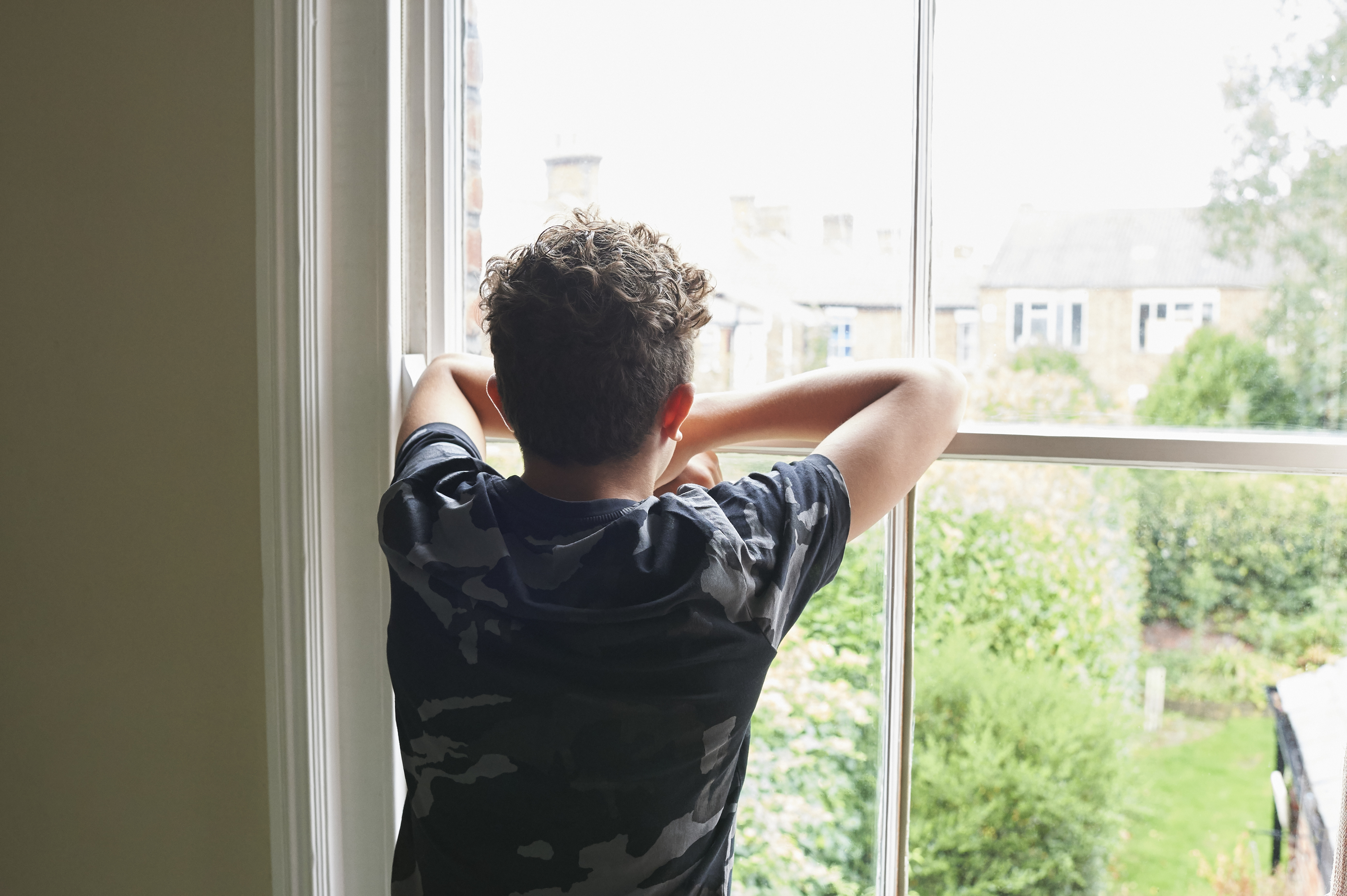 A person with curly hair looks out of a window, with arms resting on the window frame, gazing at a garden scene outside