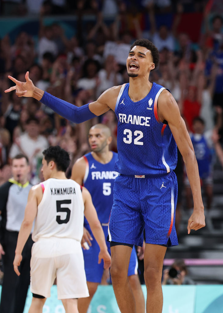Victor Wembanyama in a France basketball jersey (#32) on the court, gesturing with his hand. Yuki Kawamura (#5) in a Japan jersey is in the background