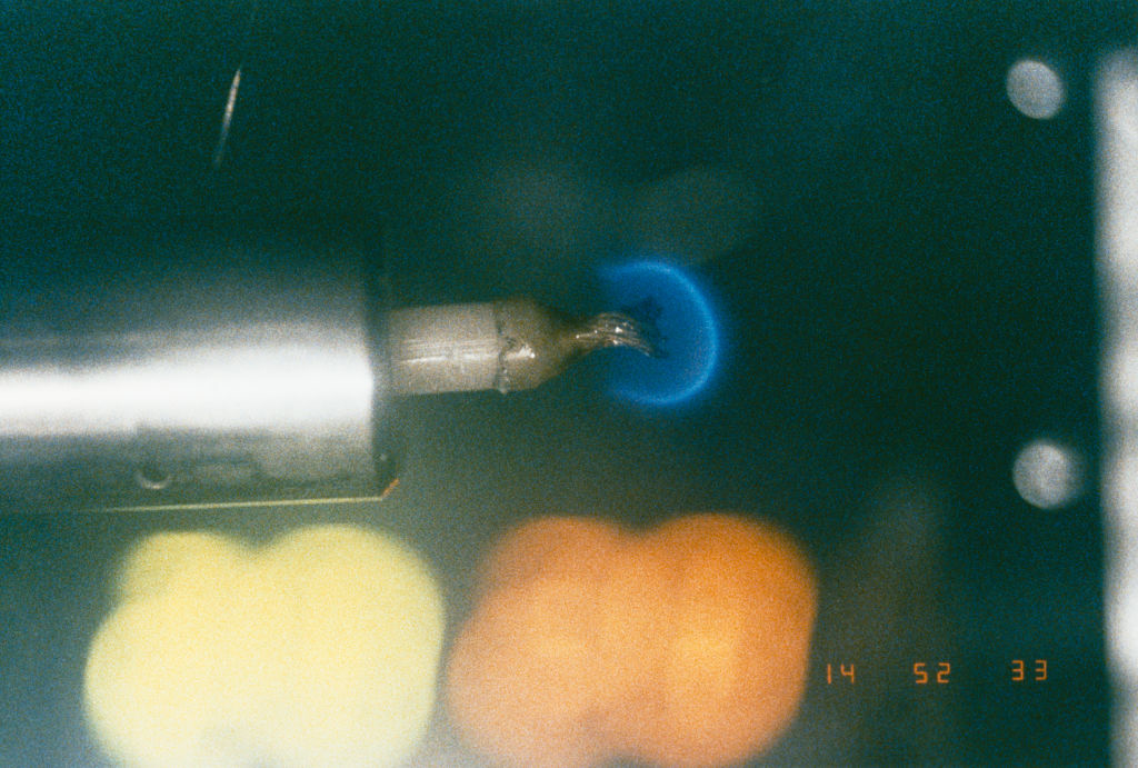 Close-up of a soldering iron tip, with a small piece of melted solder at the end, held against a blurred background of lights