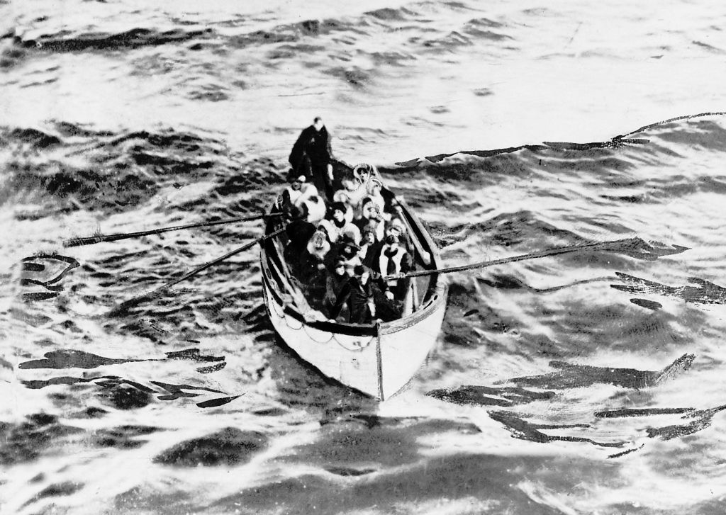 A black-and-white image showing people in a lifeboat at sea. Most are sitting while others row. The photograph appears historical