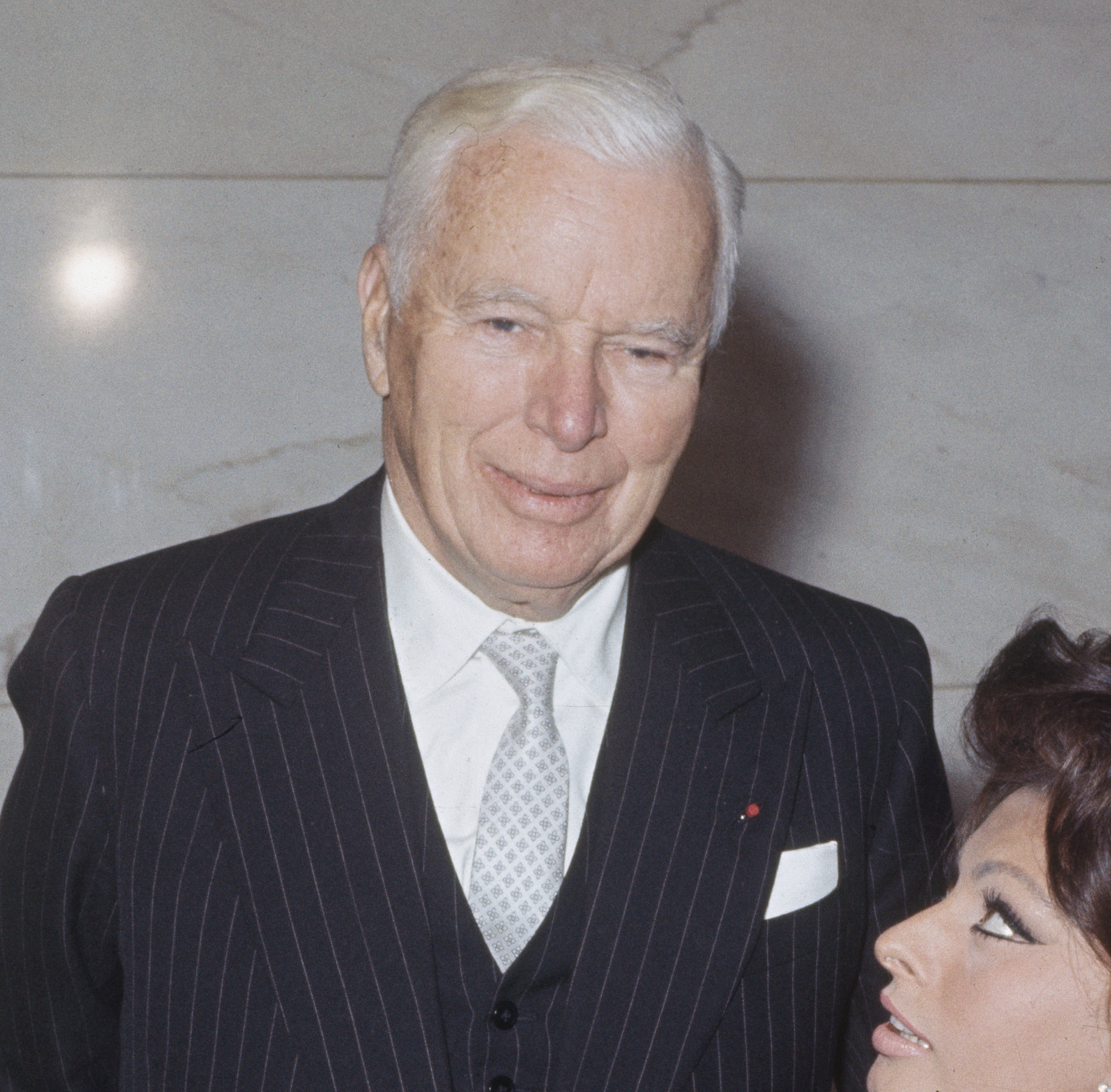 An elderly man in a suit stands next to a woman looking up at him