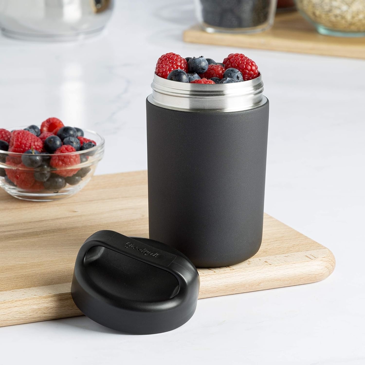 Black insulated jar filled with fresh berries and accompanied by a lid on a wooden board; a glass bowl with more berries is in the background