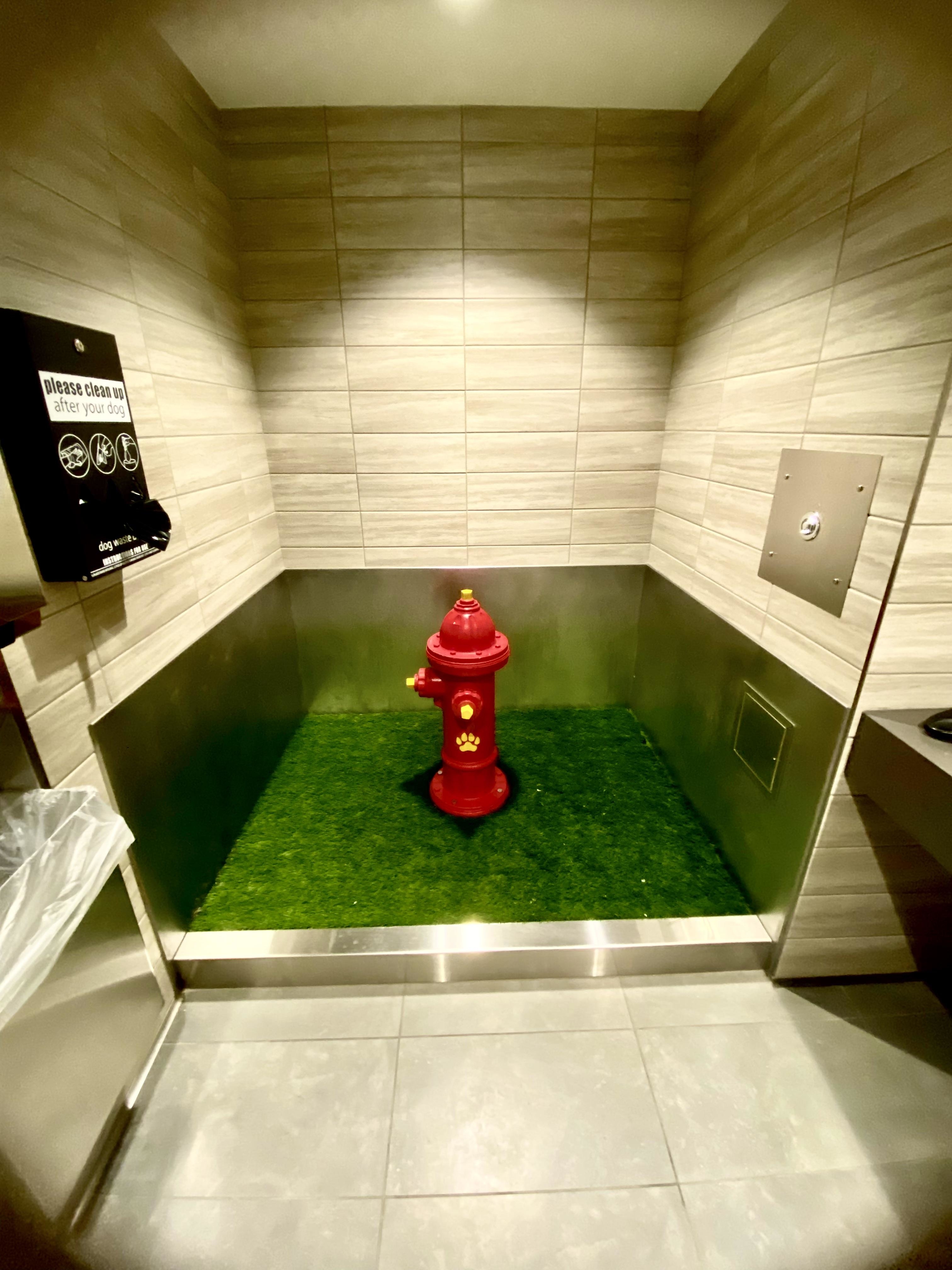 Fire hydrant-shaped object placed in the middle of a room with a fake grass patch, possibly a dog restroom