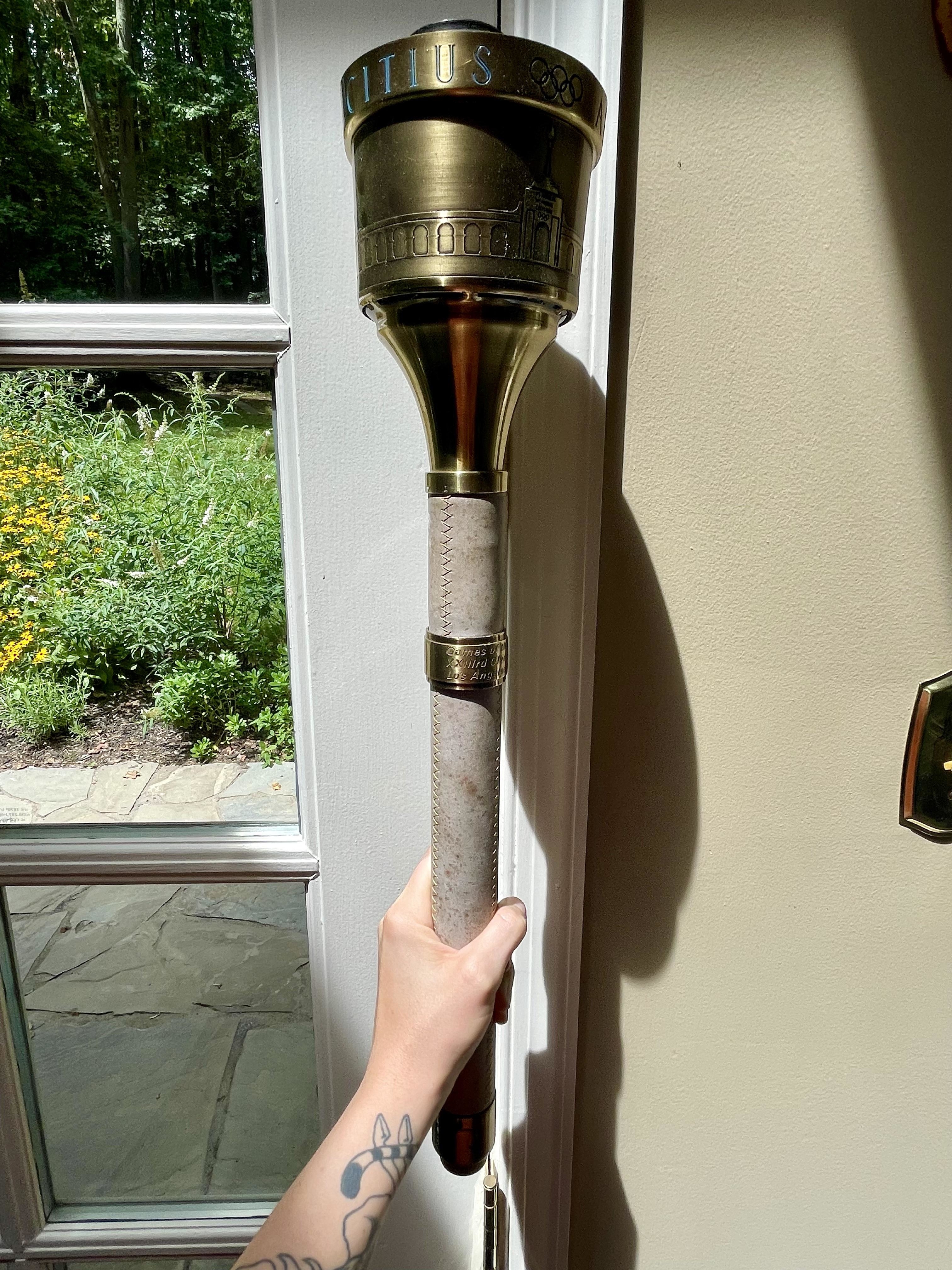 A person holds an Olympic torch near a window with a garden visible outside. The torch has a metal top with inscriptions and a stone textured handle