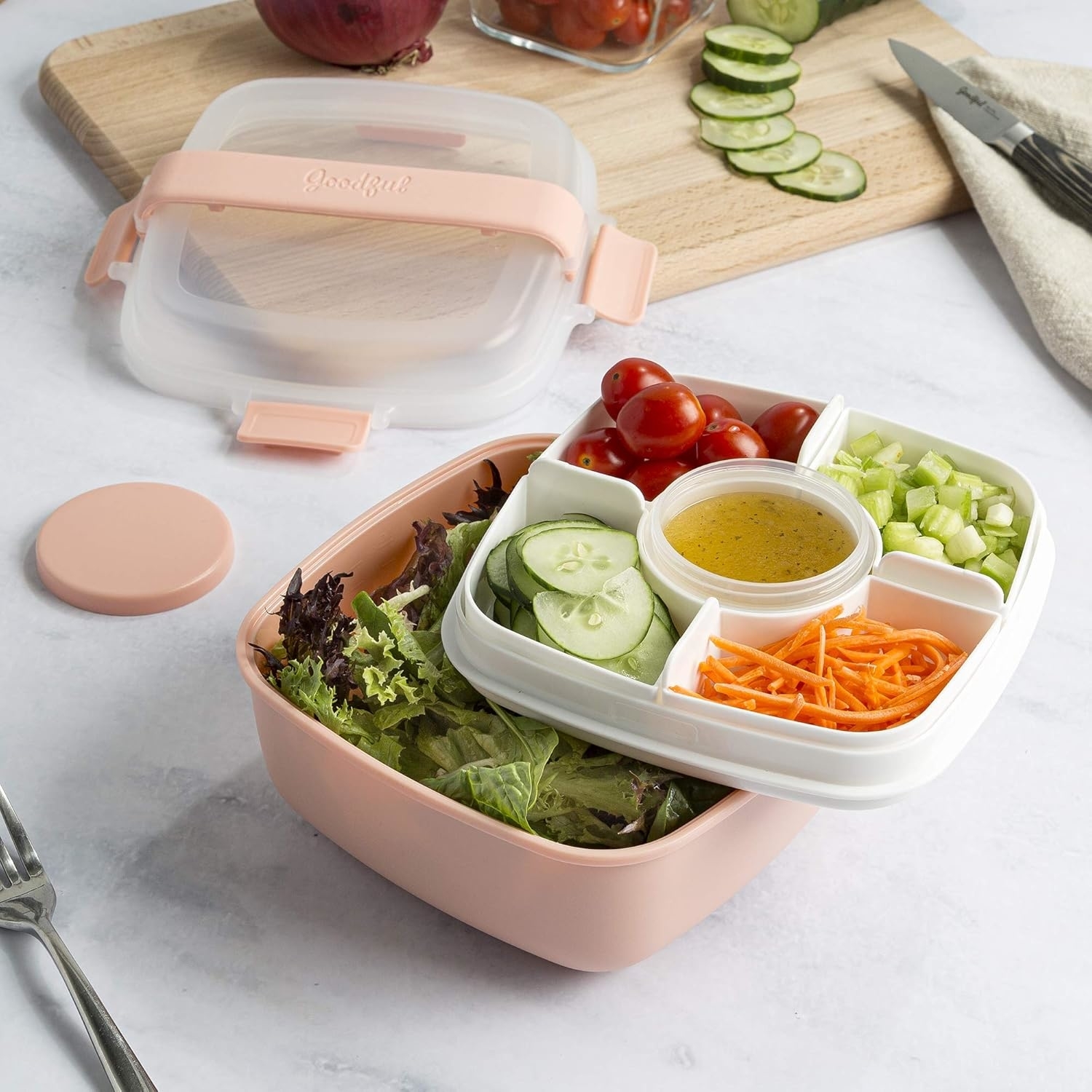 Bento-style lunch container with compartments holding cherry tomatoes, celery, sliced cucumbers, shredded carrots, mixed greens, and a central cup of dressing