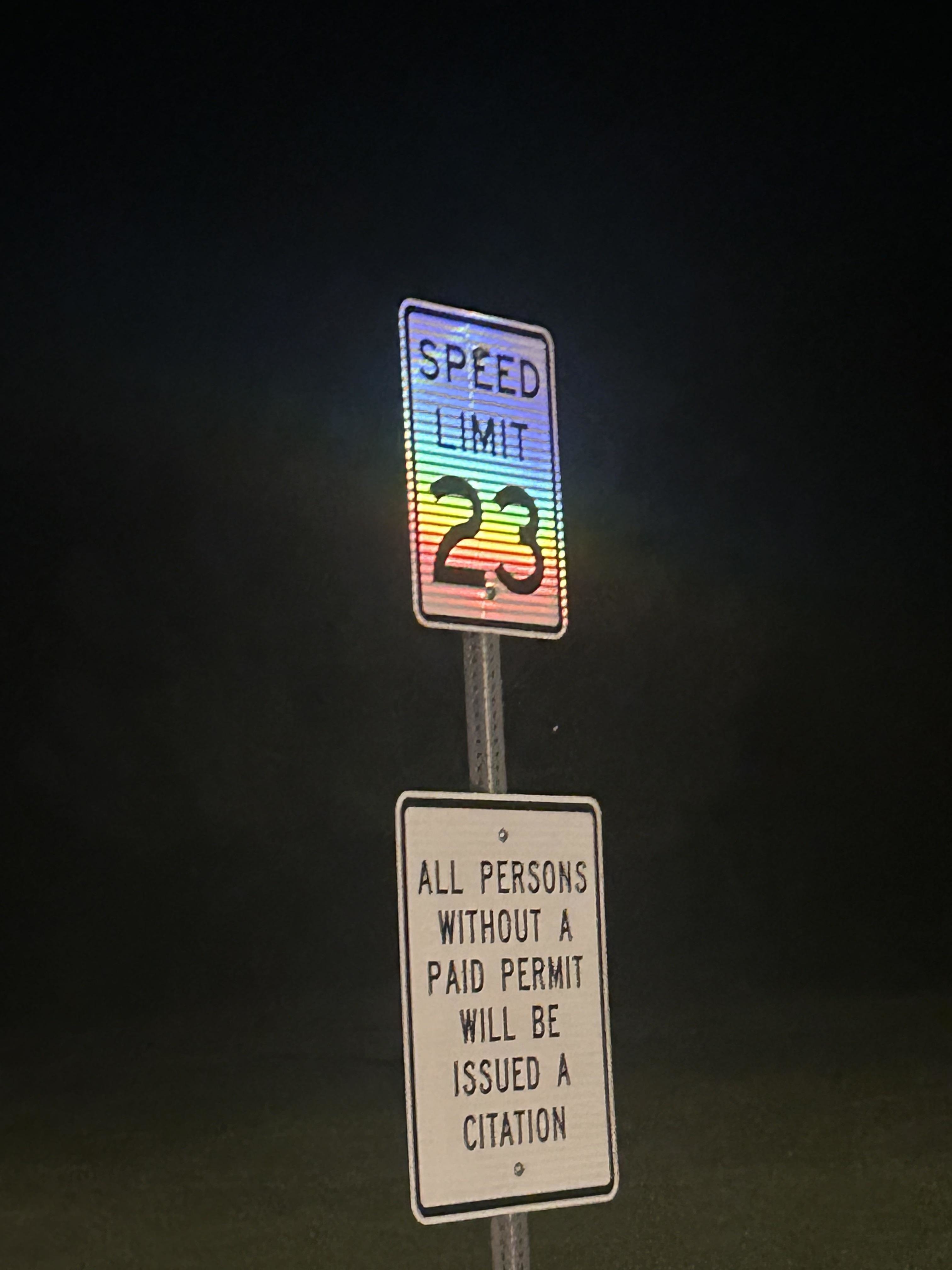 Speed limit sign reading "23" with a rainbow gradient. Below, a sign says "All persons without a paid permit will be issued a citation."