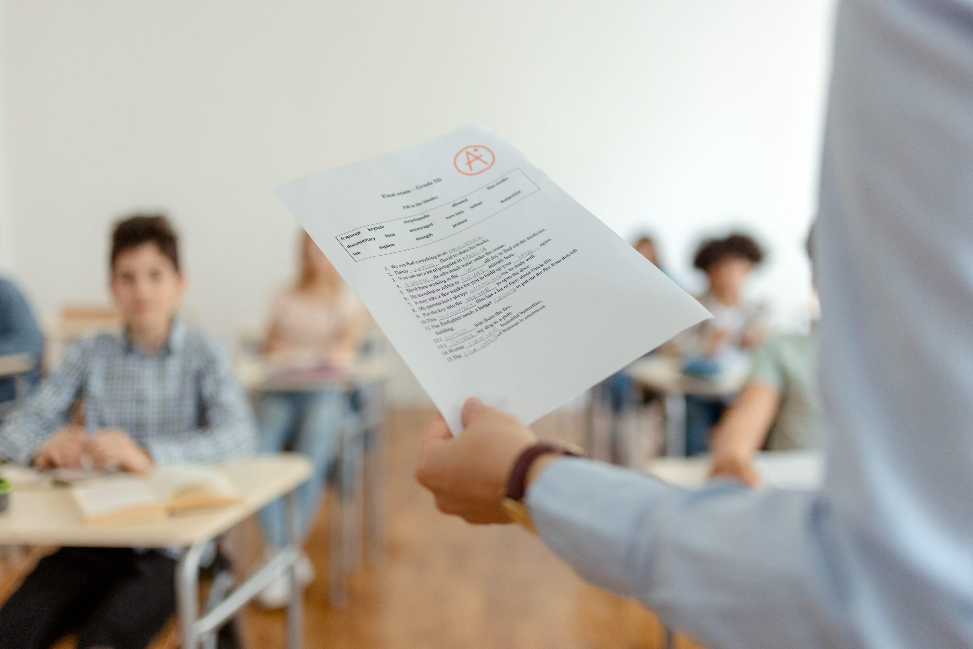 A teacher holds a test paper with an "A+" grade on it in front of students seated at desks in a classroom