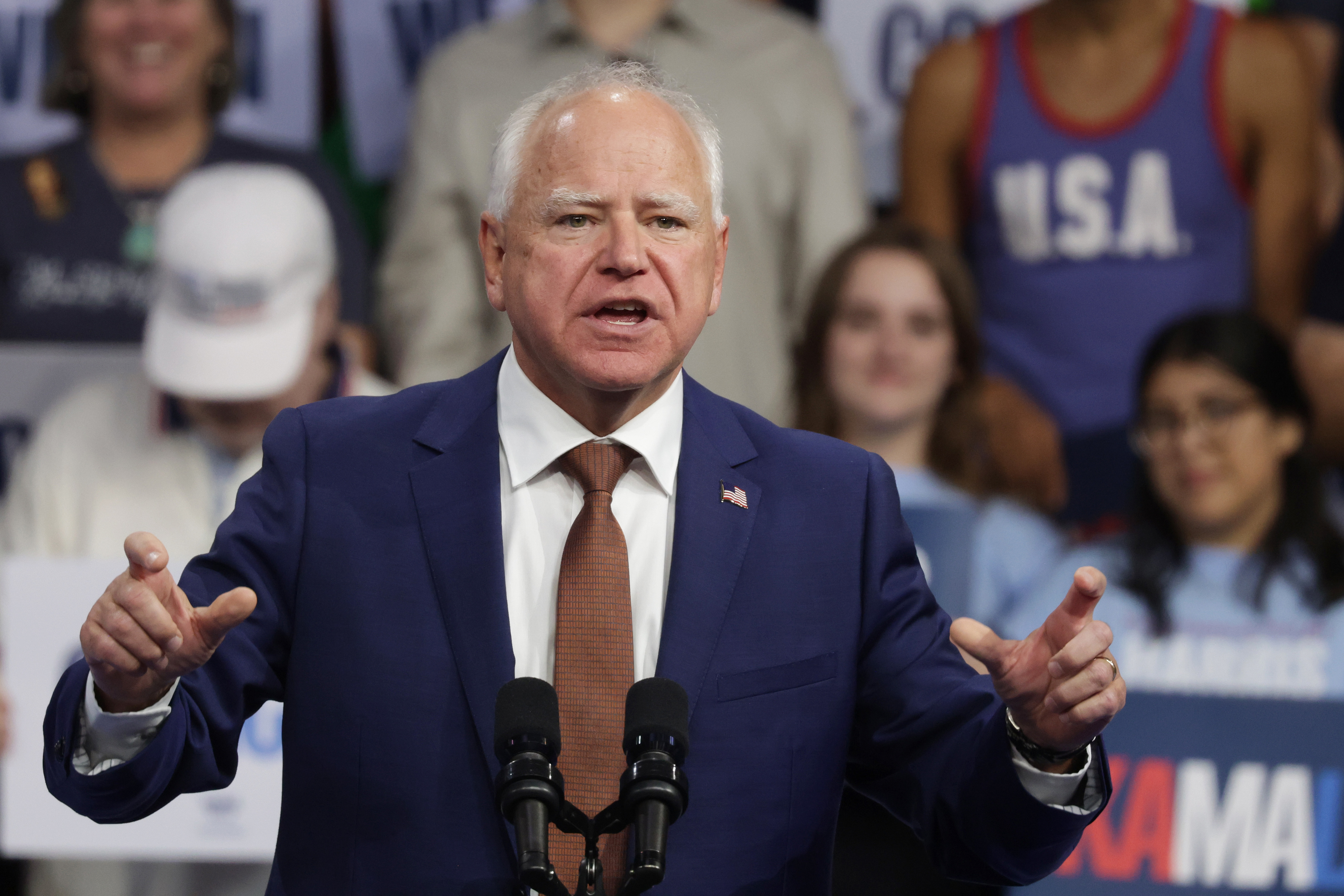 Tim Walz, wearing a blue suit and brown tie, speaks at a podium with microphones. A crowd of supporters is behind him, some holding signs