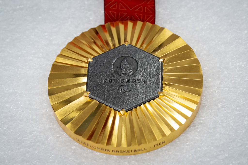 A gold Paralympic medal with "PRIS 2020" and a wheelchair basketball emblem. The medal rests on a textured white surface and is attached to a red ribbon