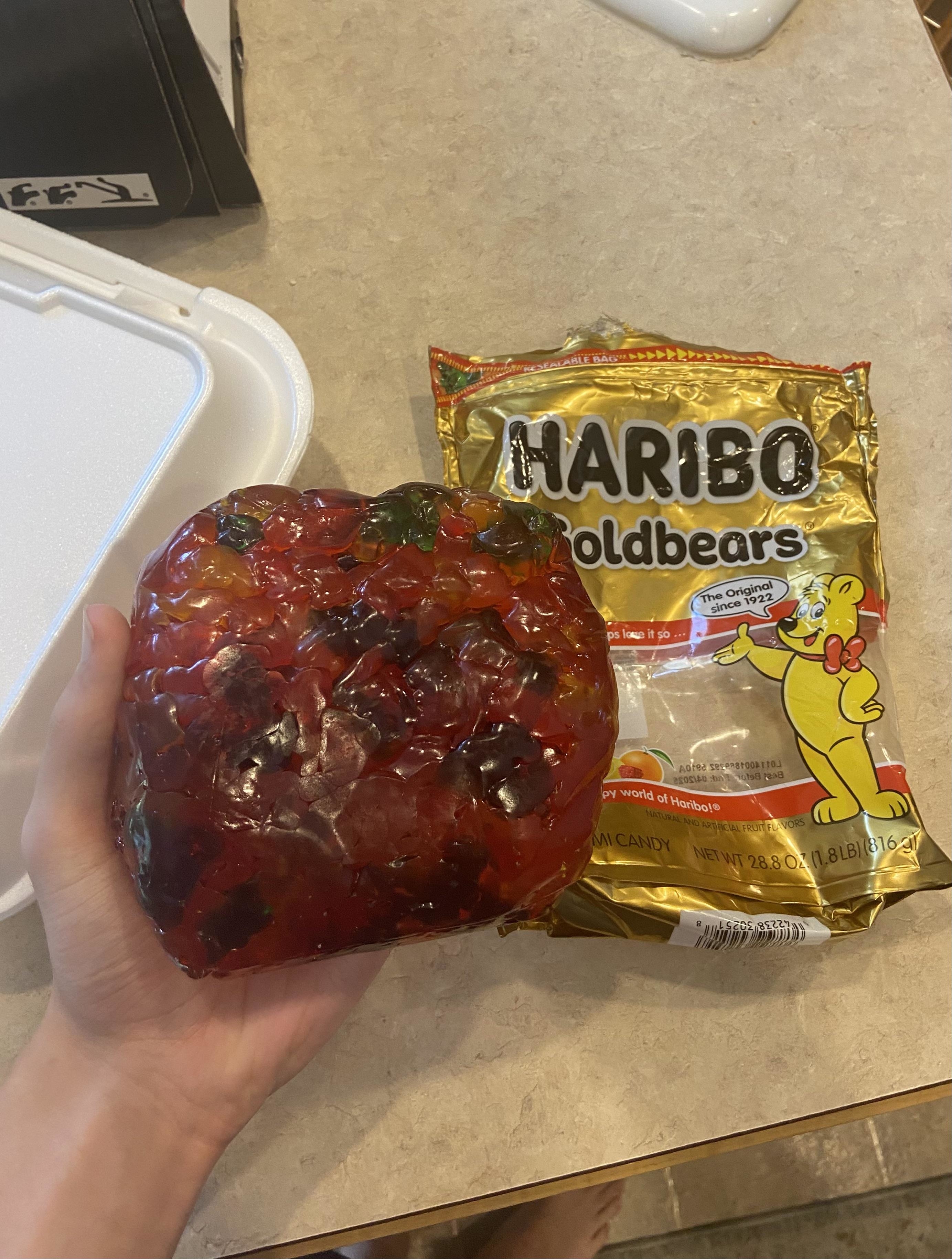 Hand holding a large, solidified mass of gummy bears next to an open bag of Haribo Goldbears on a kitchen counter