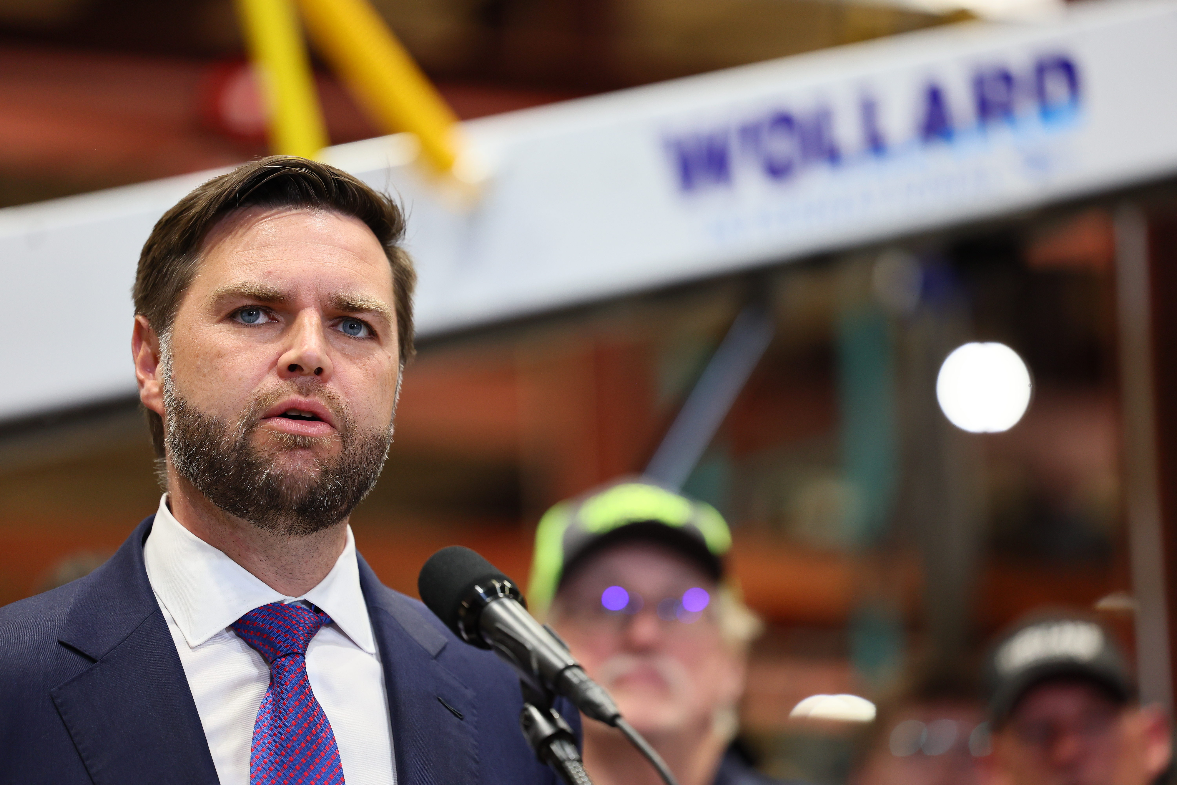 J.D. Vance speaking at a podium with a microphone, industrial setting in the background. A man in a cap is blurred behind him