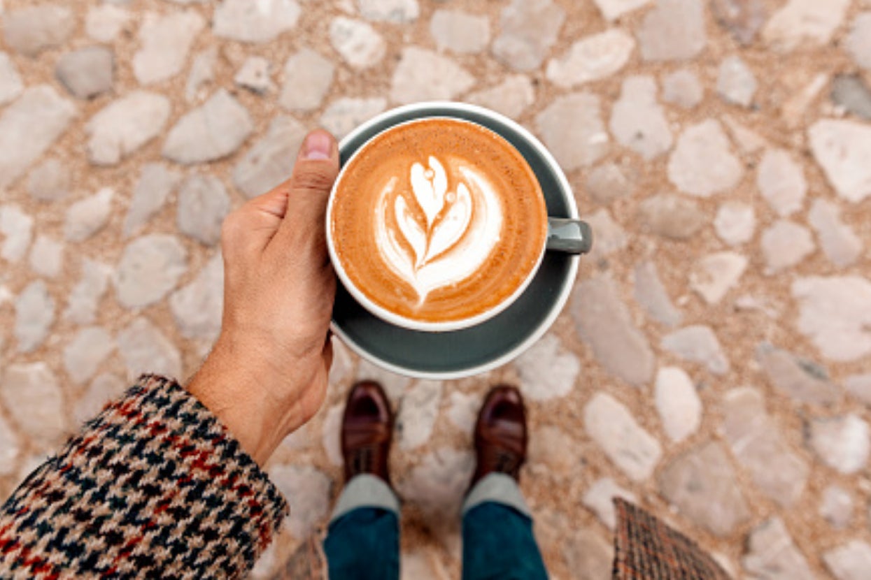 A hand holding a cup of coffee with latte art, viewed from above. The person is wearing a houndstooth coat, blue jeans, and brown shoes standing on a cobblestone pavement