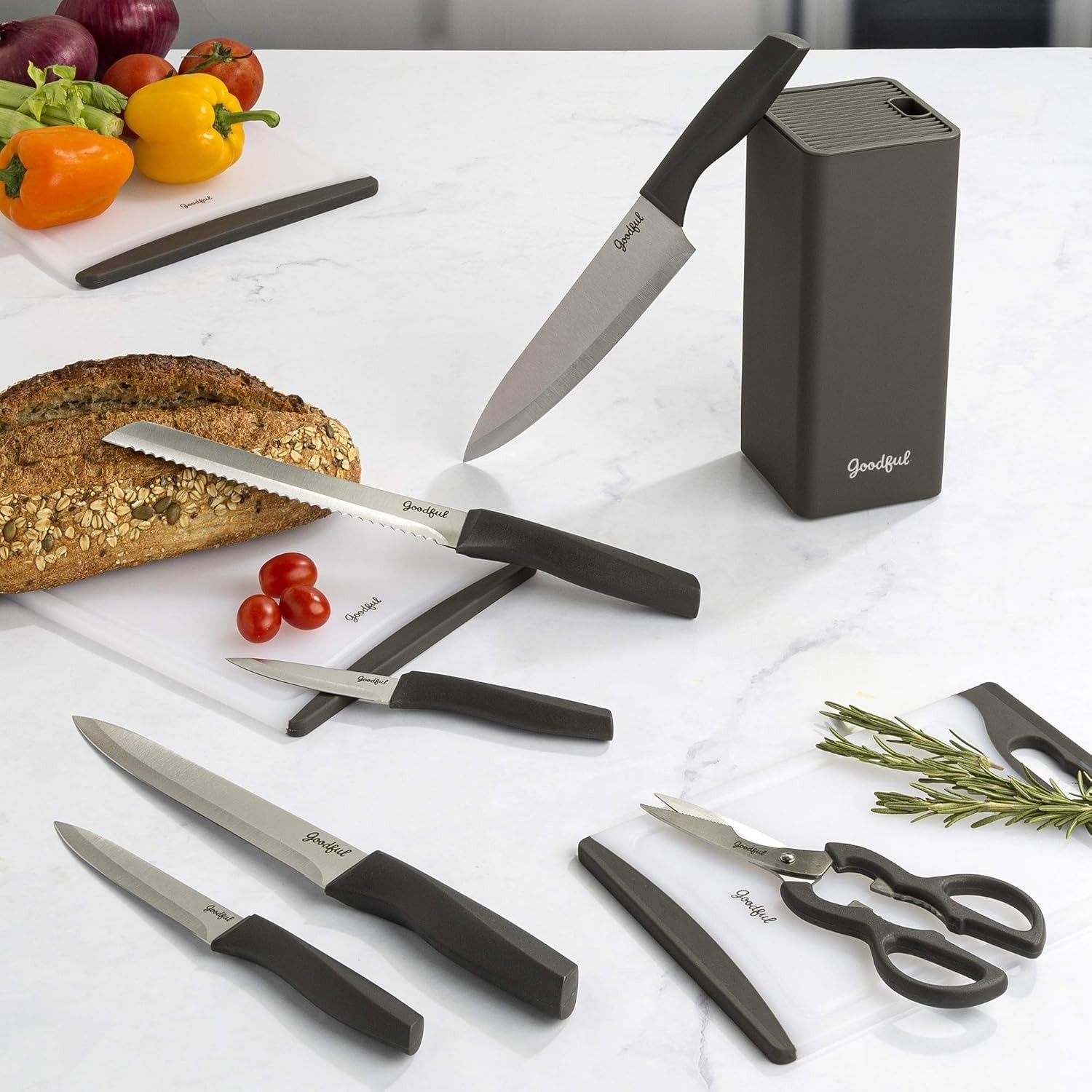 Various Goodful kitchen knives and shears, including bread and chef's knives, appear on a white marble surface with vegetables and sliced bread
