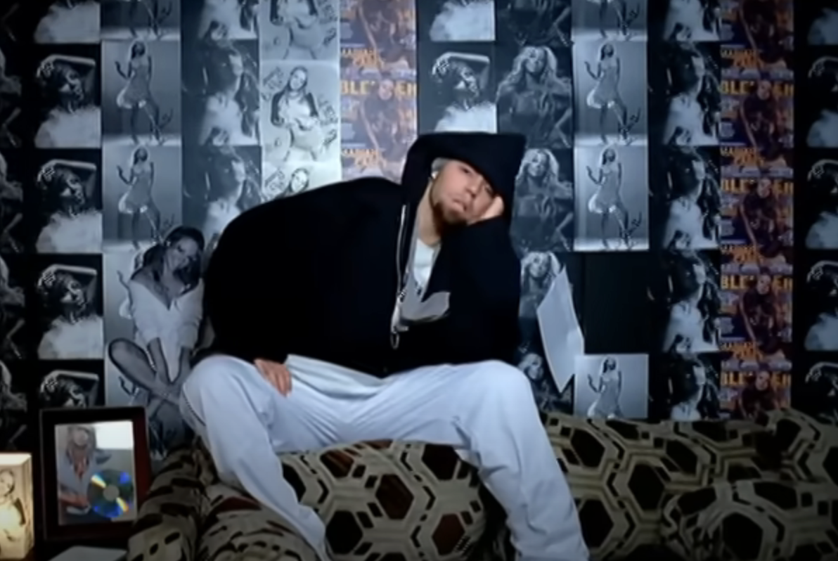 Man in a hoodie and white pants sits on a patterned couch, leaning his face on his hand in front of a collage-covered wall featuring various celebrity posters