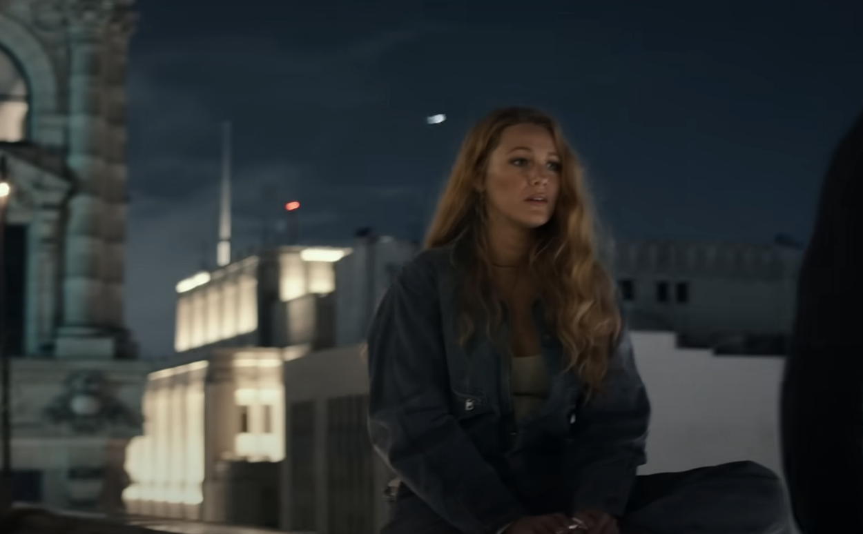 Blake Lively sits on a rooftop at night, wearing casual denim clothing, with city buildings lit in the background