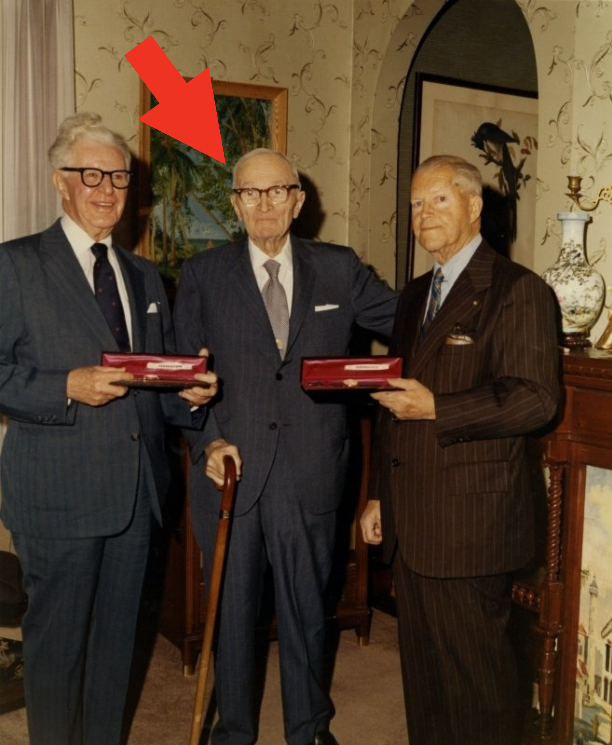 Three elderly men in suits stand together, two holding rectangular boxes. They are in a formal room with a cane near one man. Names unknown