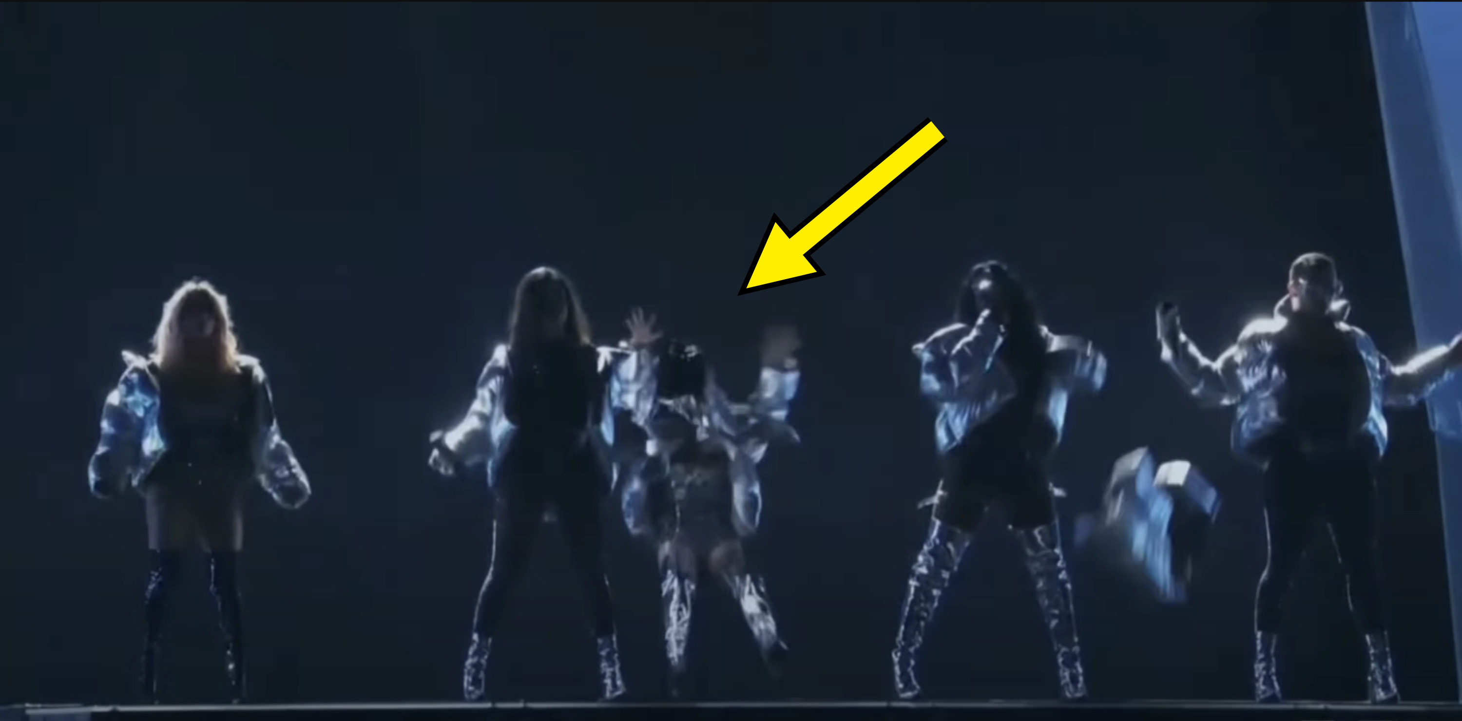 Five unidentified performers on stage in futuristic outfits and shiny boots, striking dynamic poses during a performance under dim lighting